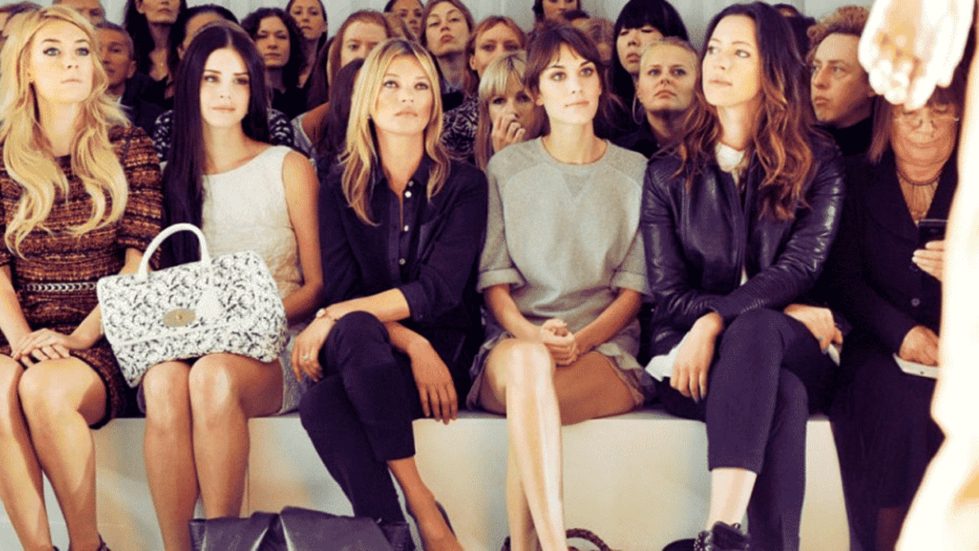 A group of women seated on a bench, attentively watching a fashion show in a stylish venue.