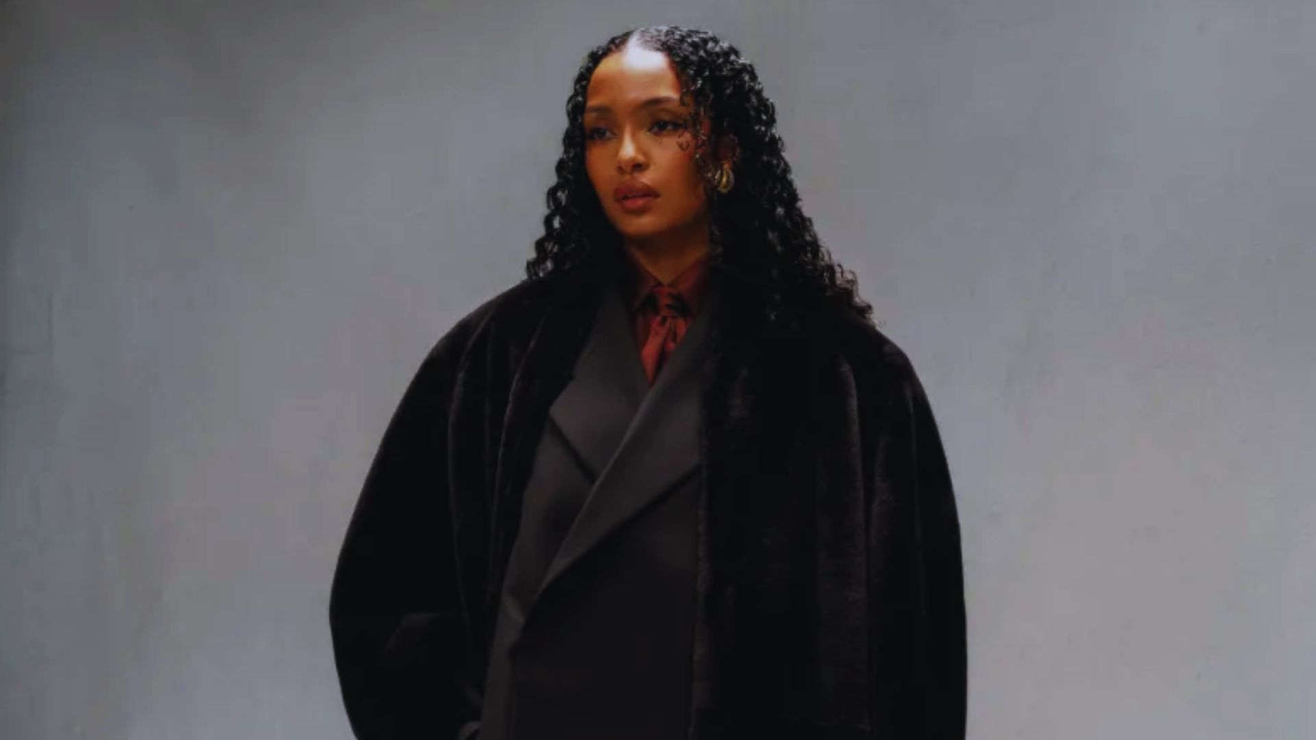 A woman dressed in a black coat and red shirt poses, highlighting her fashionable ensemble.