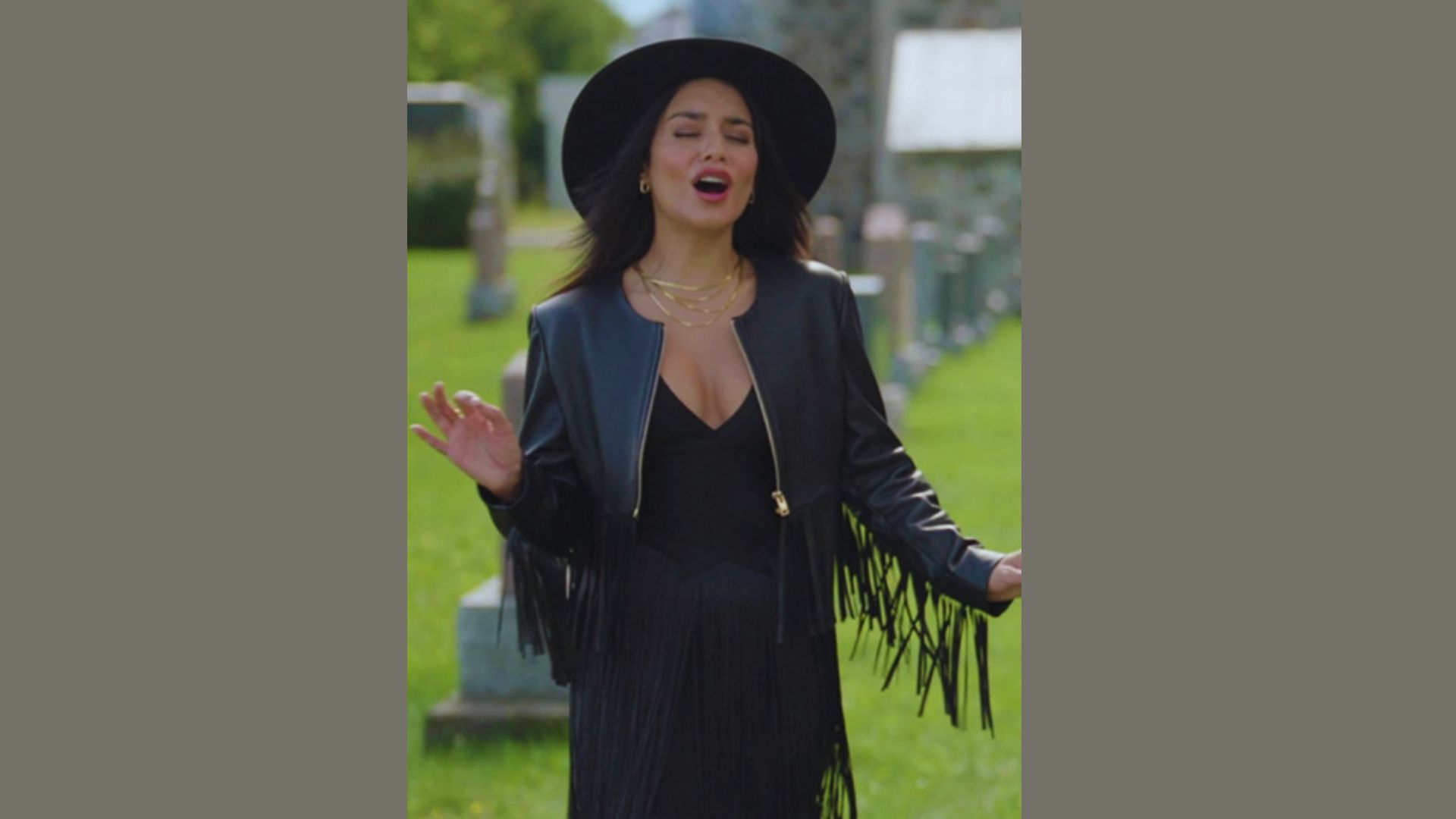A woman wearing a black dress and hat, singing energetically during a live performance.