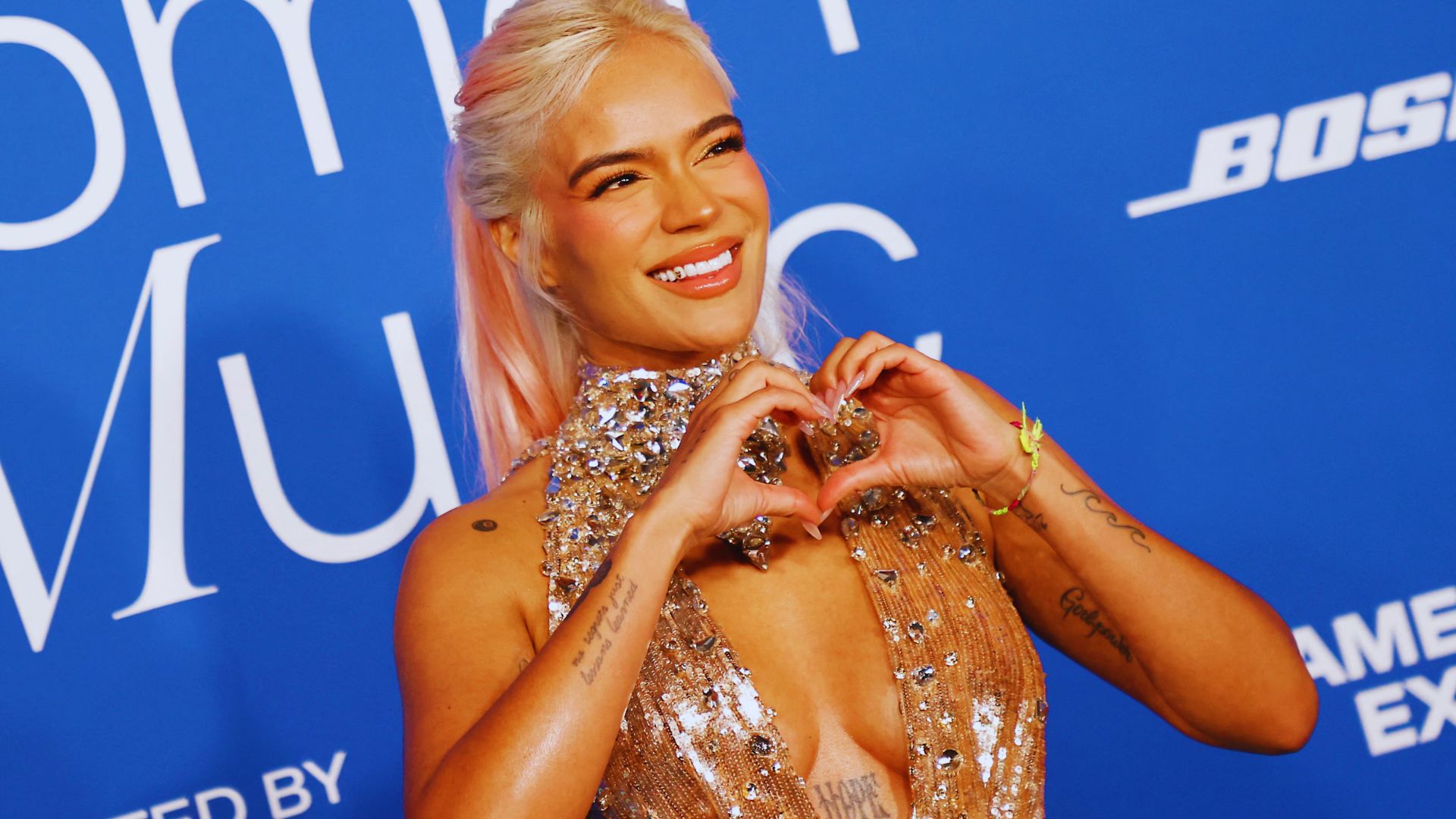 A woman with pink hair and tattoos forms a heart shape with her hands, smiling warmly at the camera.