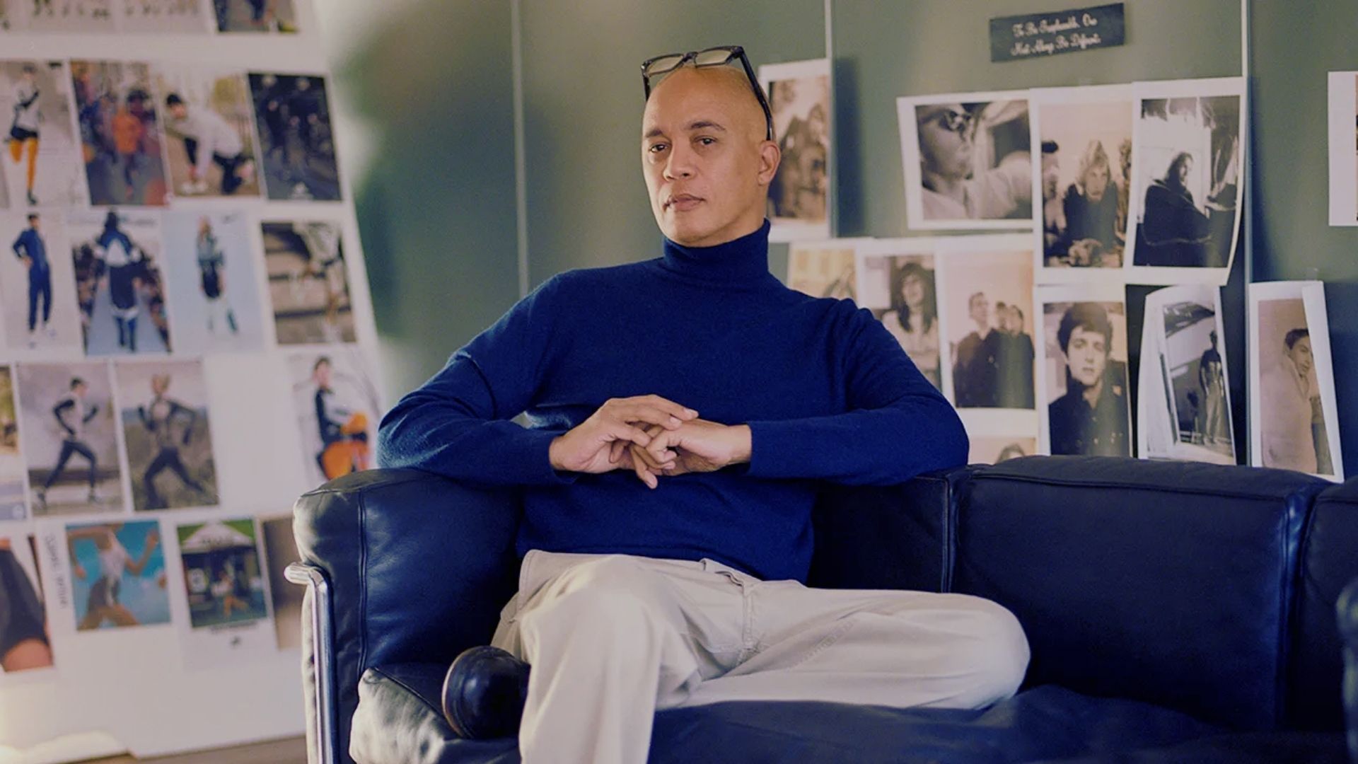 A man sits on a couch, surrounded by a wall adorned with various pictures, pondering Jennifer Aniston's wedding dress designer.