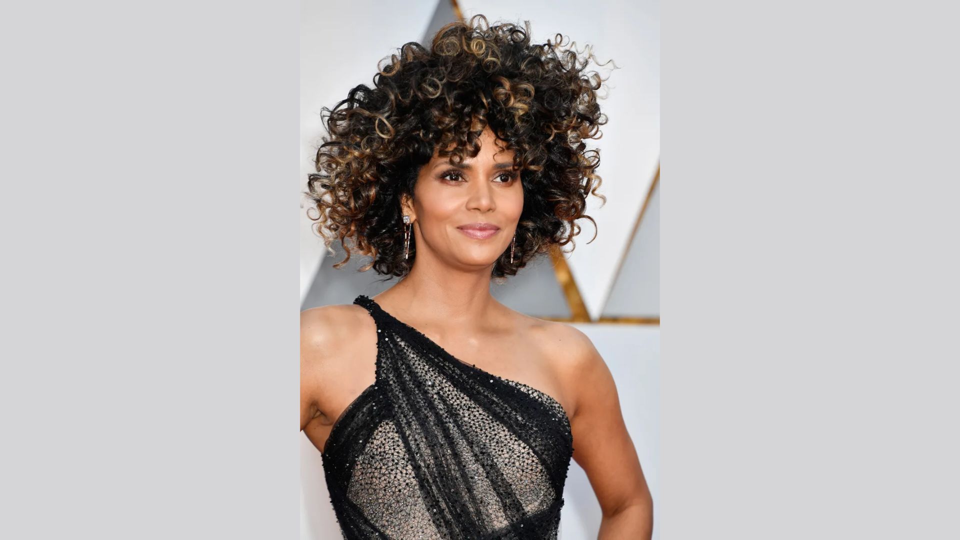 Halle Berry showcases stunning curly hair in an asymmetrical, one-shoulder style, transforming her look dramatically.