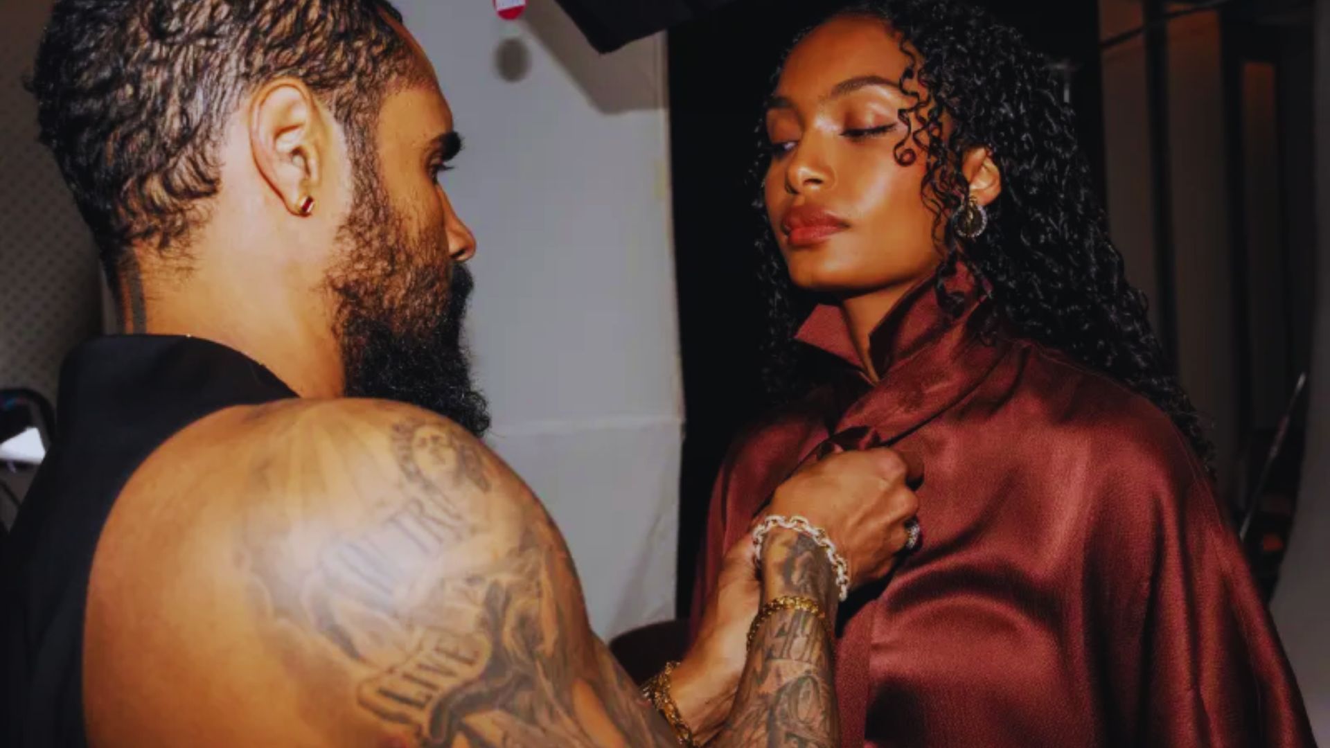 A tattooed man adjusts the shirt of a woman, focusing on her attire with a gentle expression.
