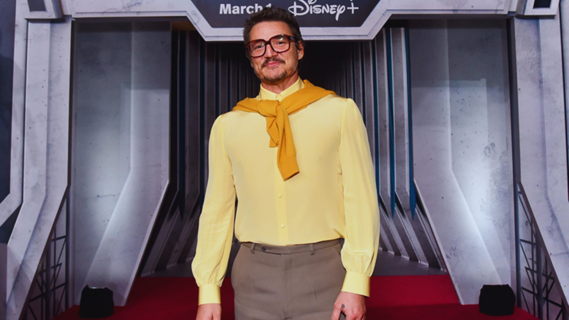 A man wearing a yellow shirt and glasses stands confidently on a vibrant red carpet.