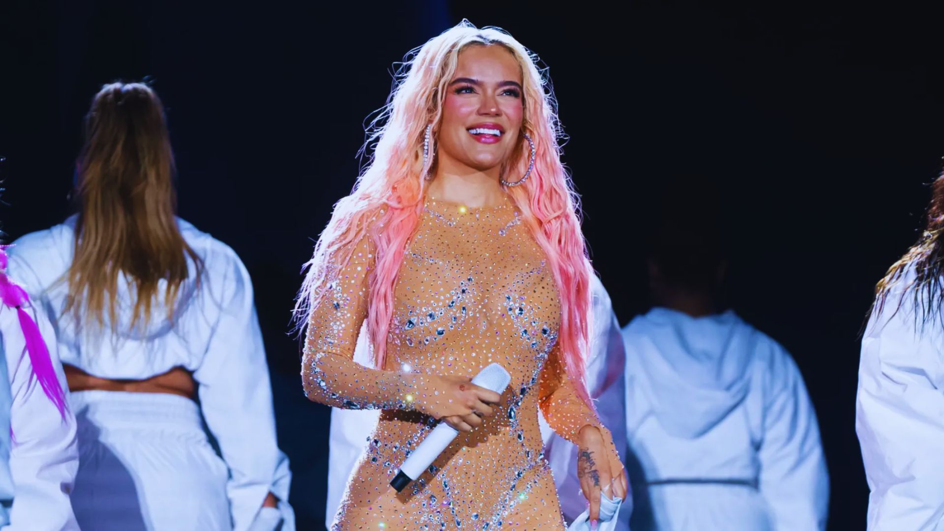 A woman with pink hair stands on stage holding a microphone, engaging the audience with her performanc