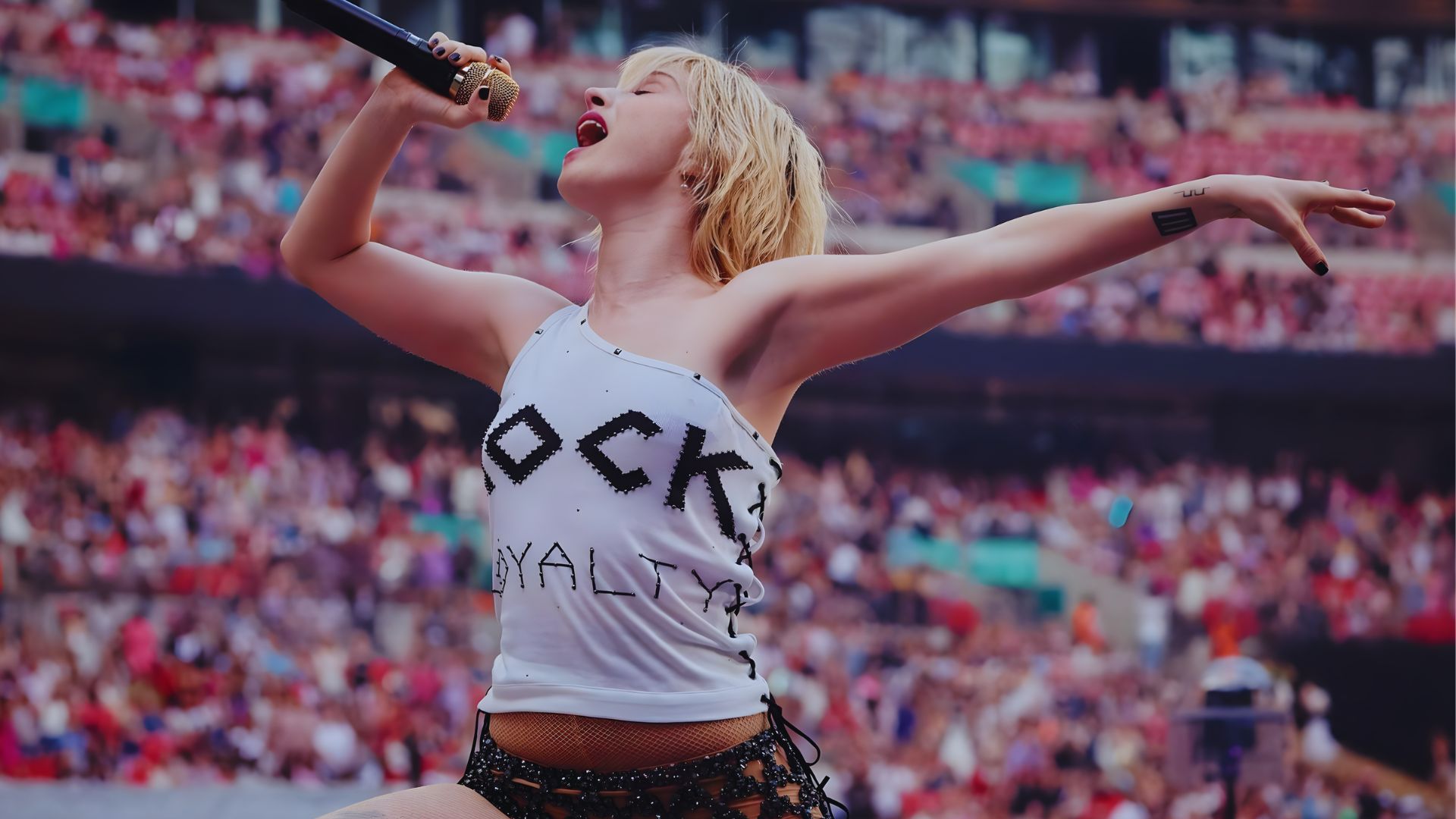 A woman in a white shirt and black shorts passionately sings into a microphone on stage.