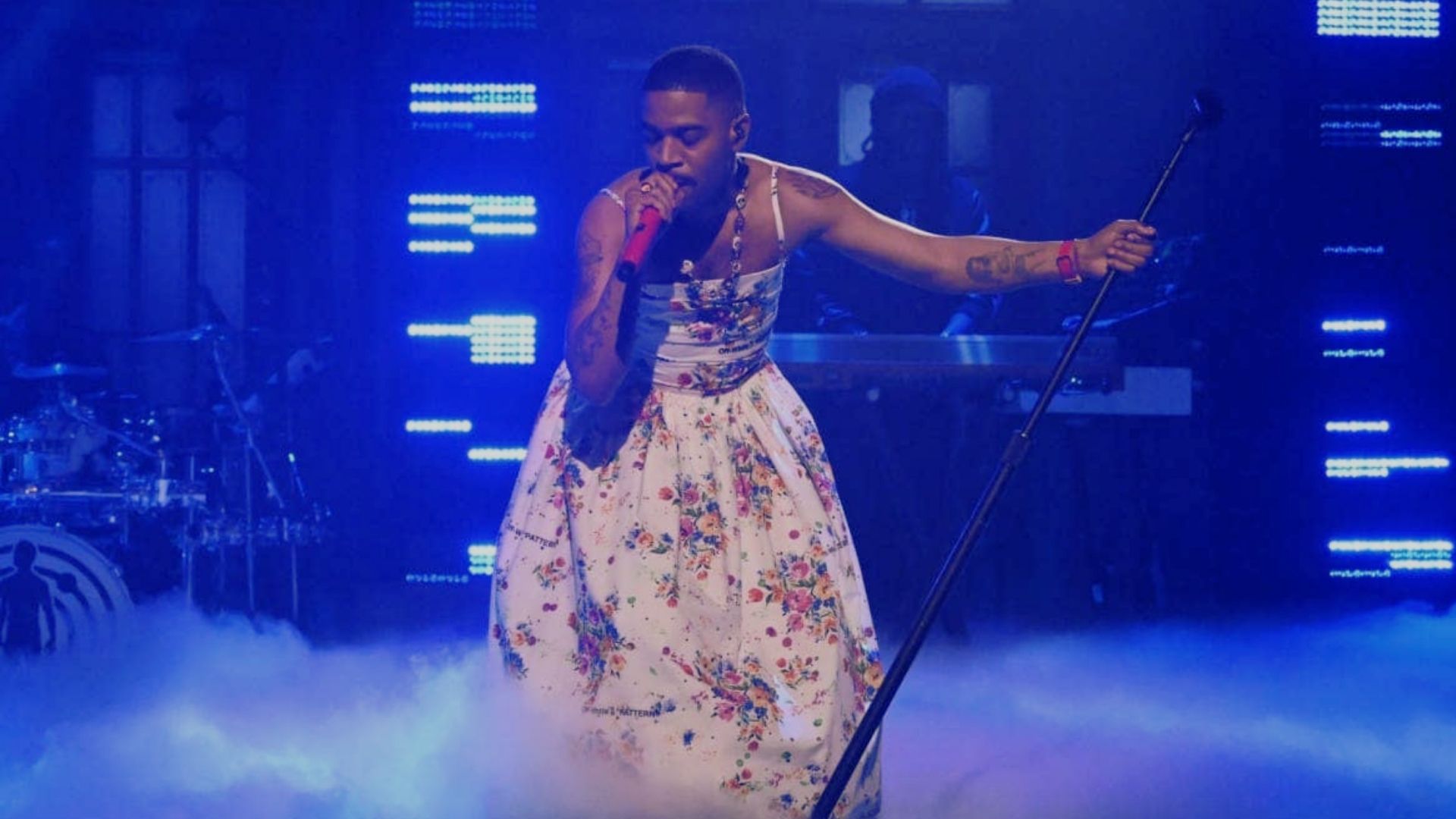 A woman in a floral dress sings passionately on stage during a performance.