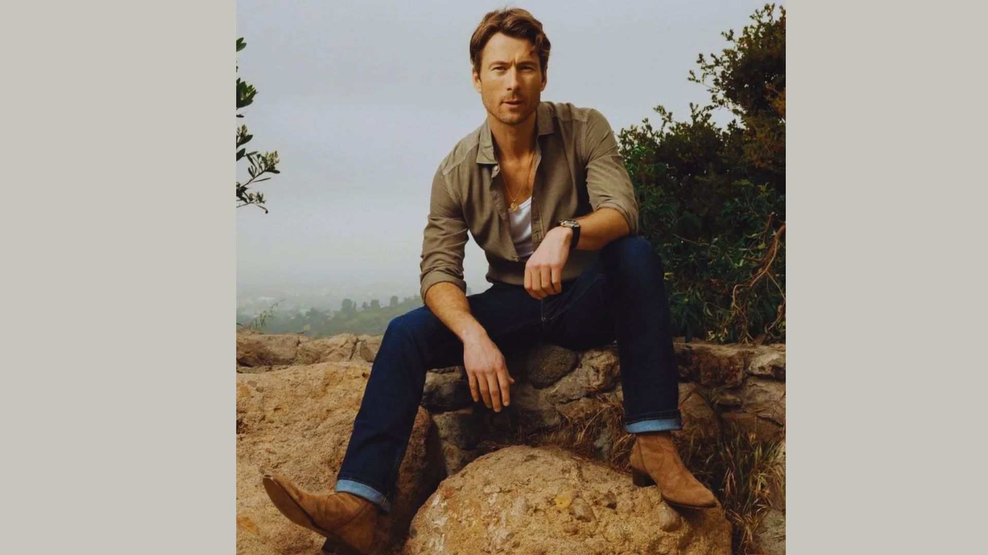 Glen Powell sitting on a rock wearing brown shirt and brown boots.