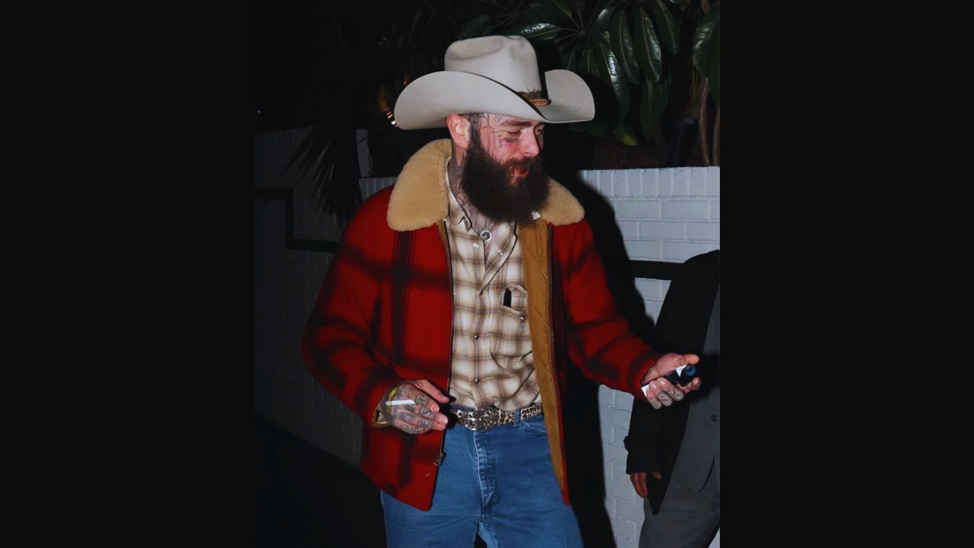 A bearded man dressed in a plaid jacket and jeans stands confidently, showcasing a casual yet stylish look.