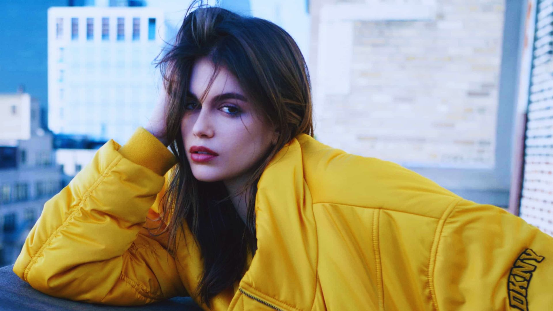 A woman wearing a yellow jacket sits on a ledge, looking thoughtfully into the distance.