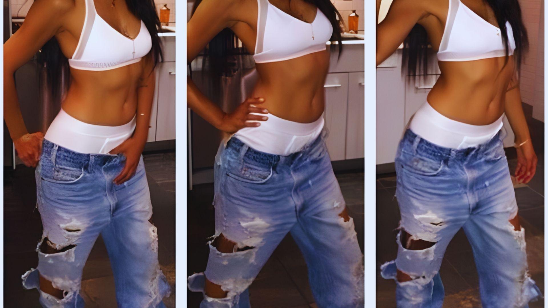 A woman in a white sport bralette and distressed jeans poses confidently in a kitchen. Her long hair cascades down, enhancing a relaxed, bold vibe.