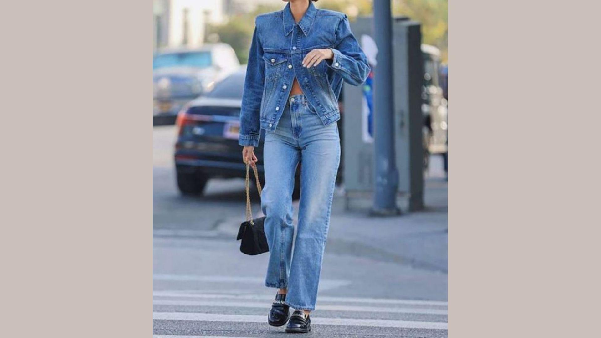 Person dressed in a denim jacket and jeans, standing casually against a neutral background.