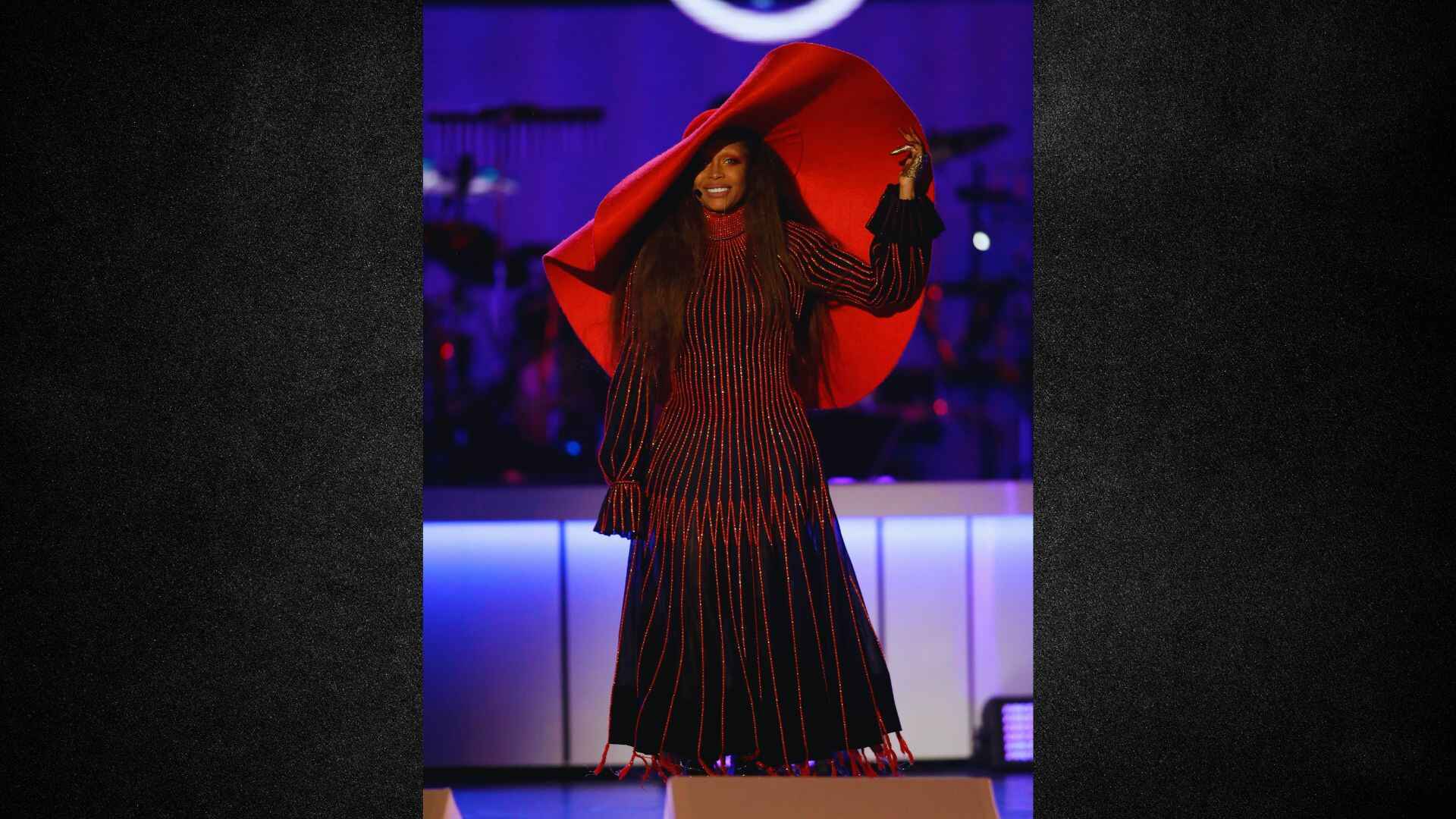A woman wearing a red dress and a black hat stands confidently, showcasing her stylish outfit.