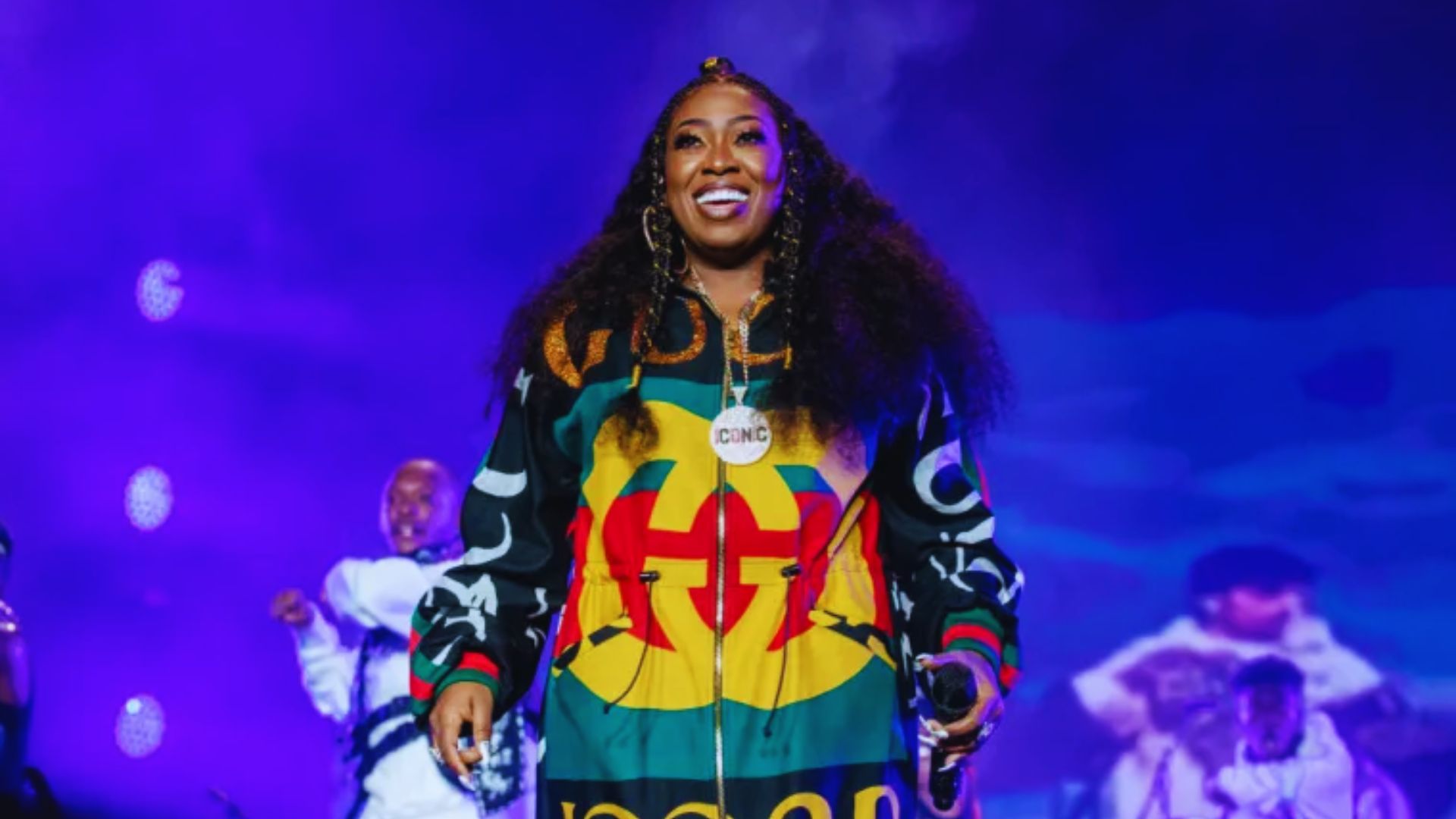 A woman wearing a vibrant, colorful jacket stands confidently on stage, engaging with the audience.