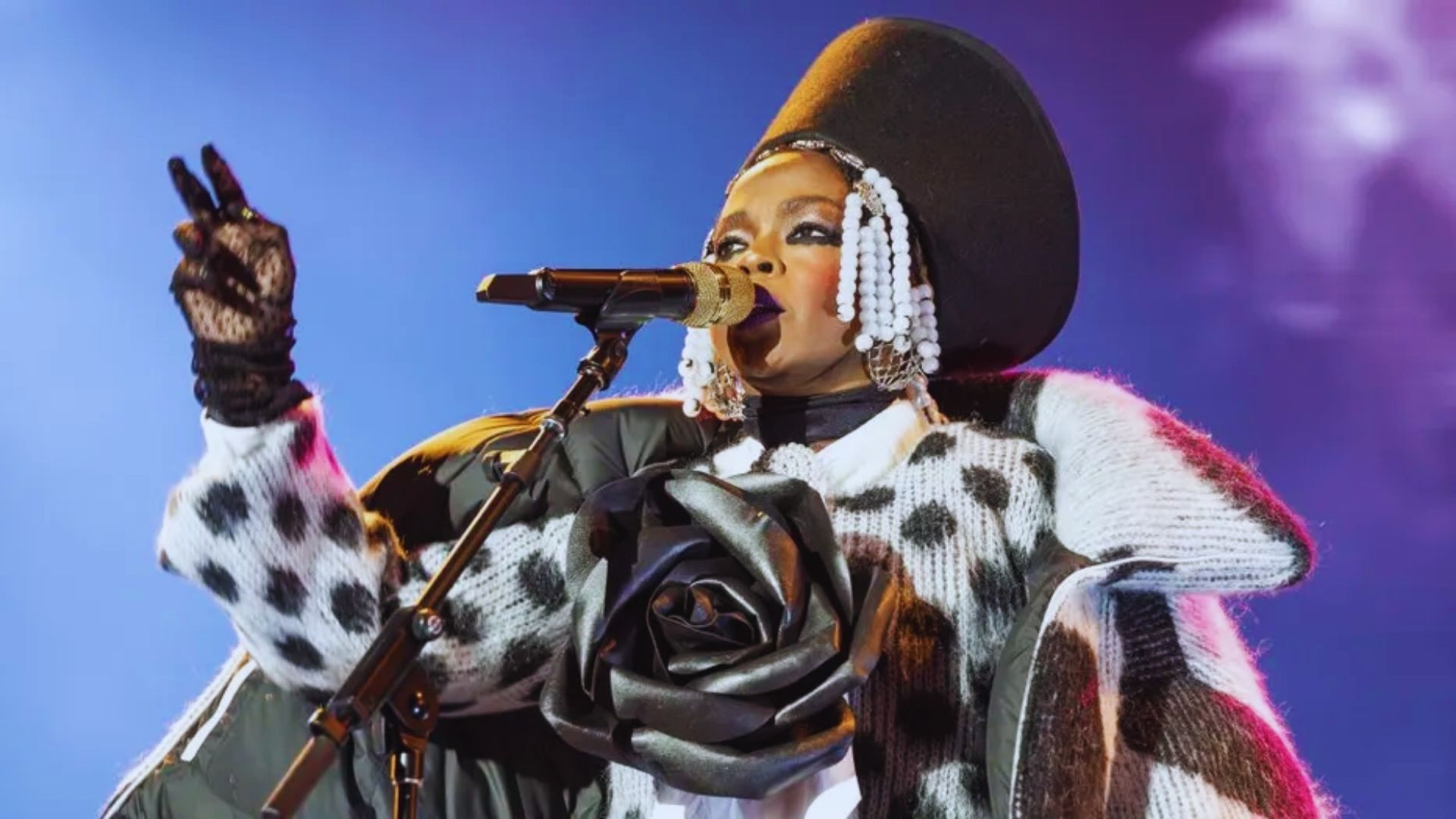 A woman wearing a fur coat and hat performs vocally into a microphone, showcasing her talent.