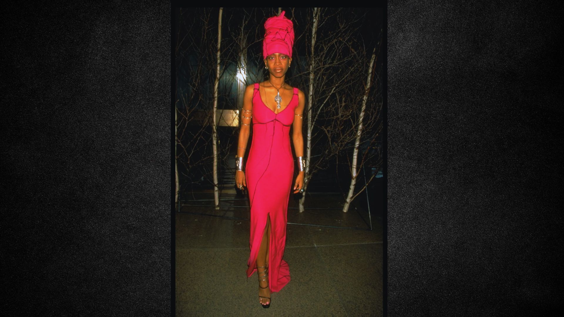 A woman wearing a pink dress and a turban, standing confidently with a serene expression.