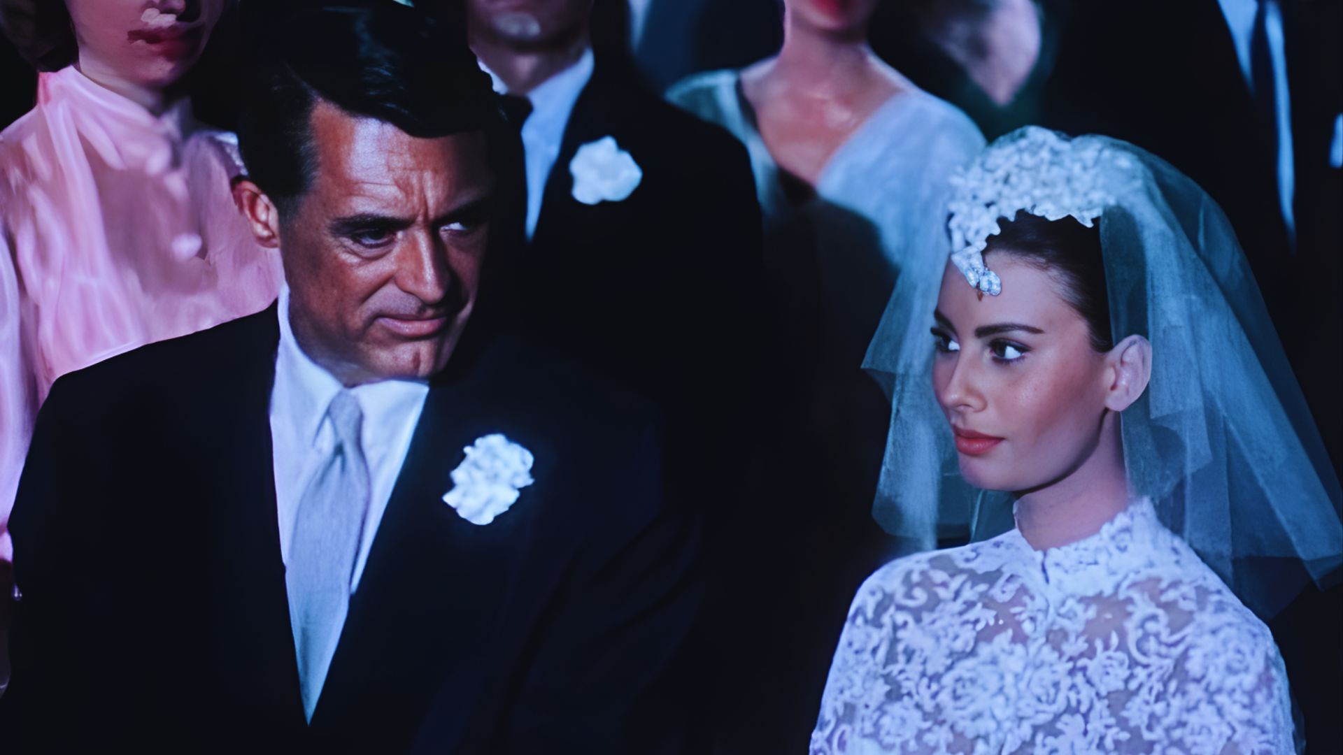 A man and woman in elegant wedding attire stand side by side, smiling at the camera in a romantic outdoor setting.