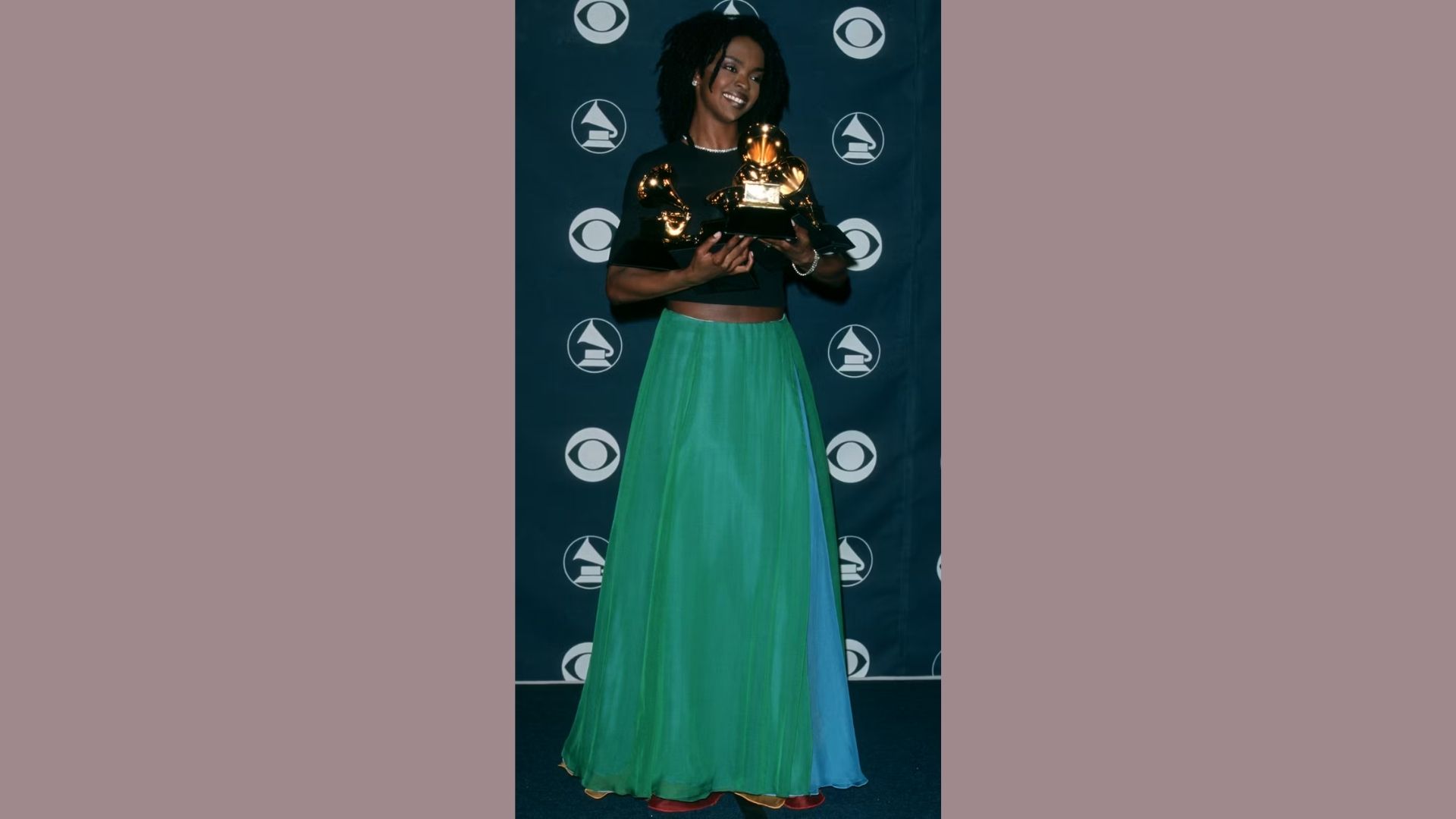 A woman in a green skirt and black top proudly holds an award, smiling at the camera.