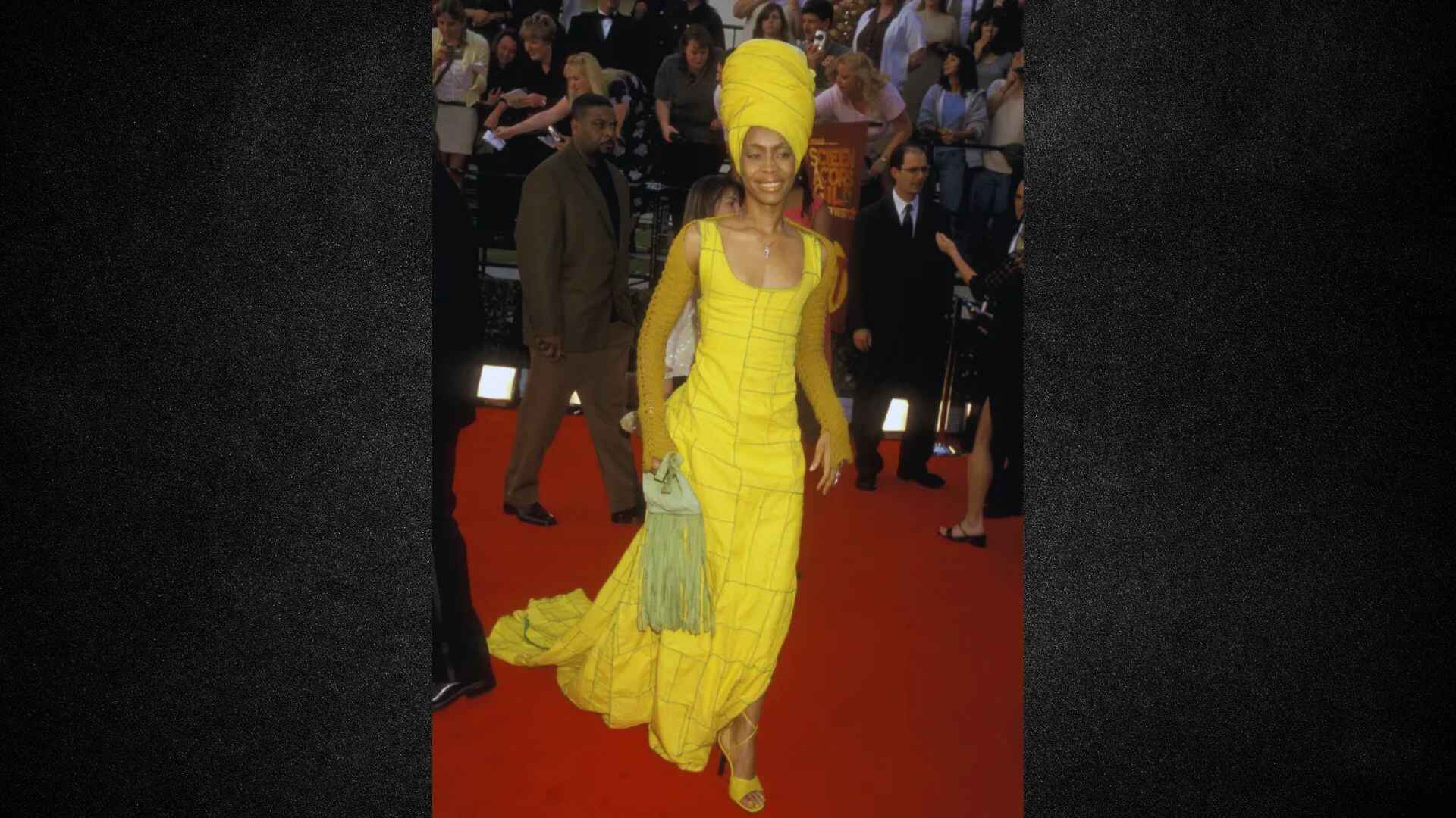 A woman in a yellow dress and turban poses elegantly on a red carpet, showcasing a vibrant and stylish appearance.