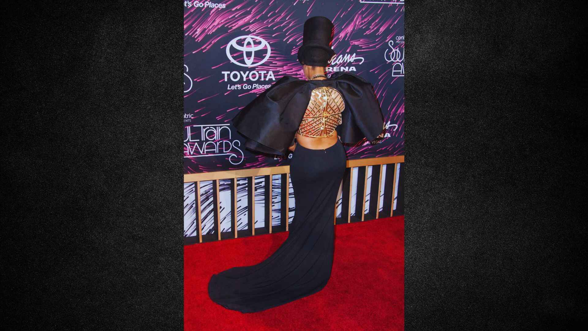 A woman in a black dress and hat poses elegantly on the red carpet at a glamorous event.