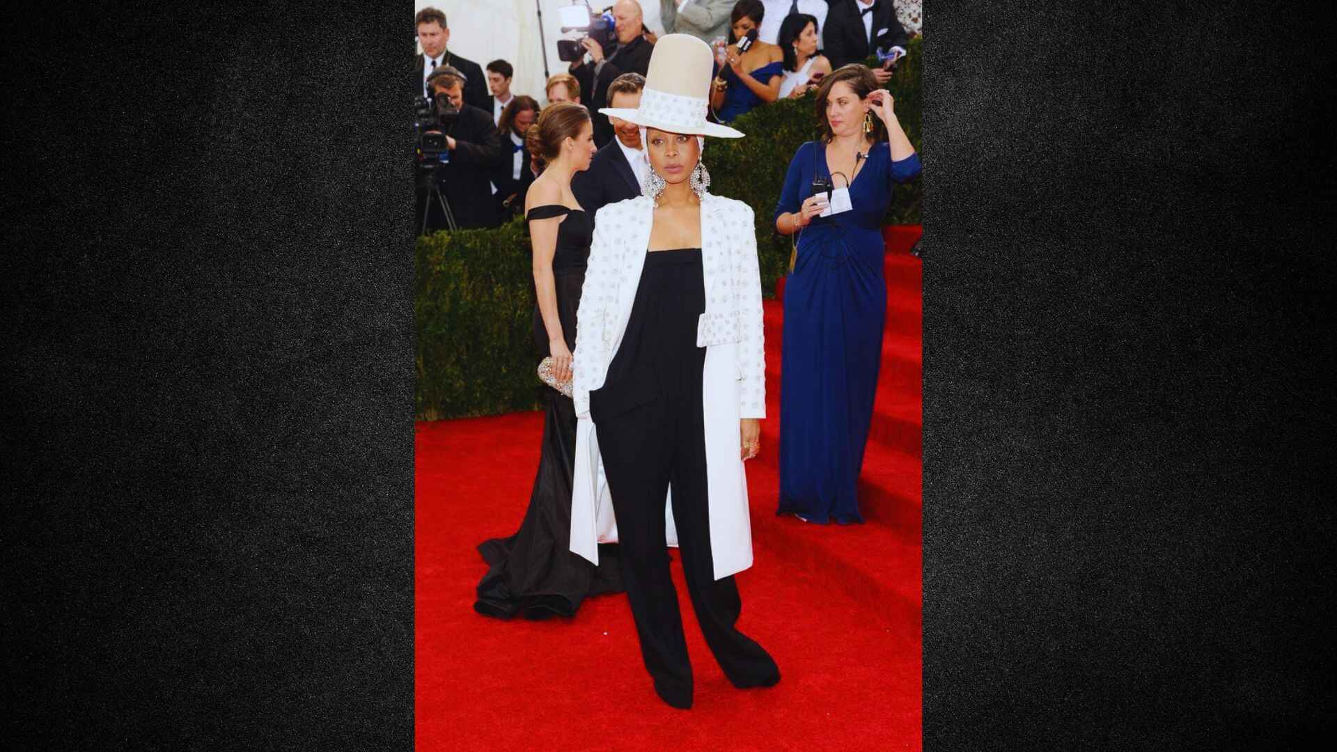 A woman wearing a white hat and black suit poses on a red carpet, showcasing elegance and style at a formal event.