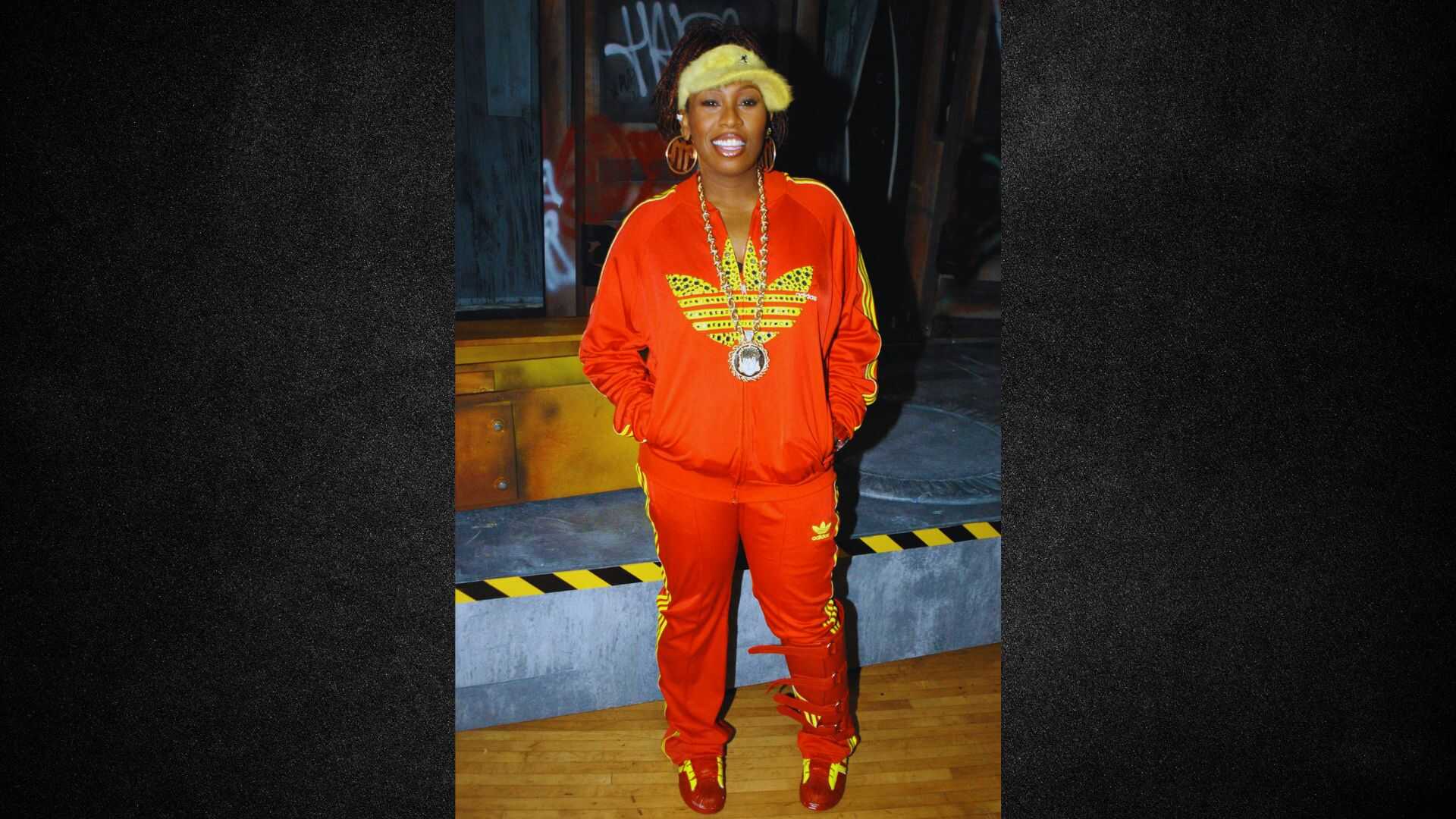 A man wearing a vibrant red suit and shiny gold shoes stands confidently, showcasing a bold fashion choice.
