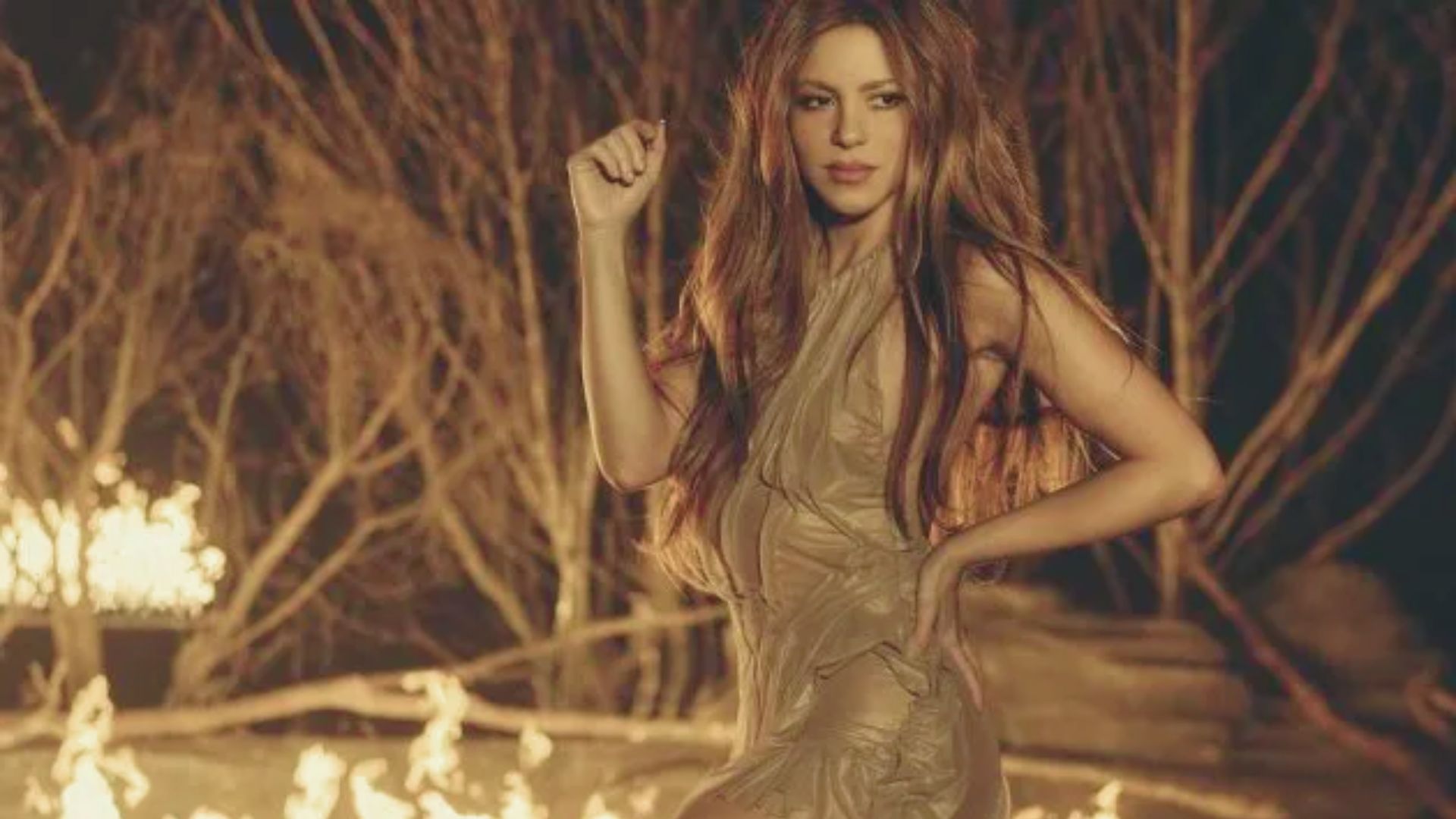 A woman in a shimmering gold dress stands confidently in front of a blazing fire, creating a striking visual contrast.