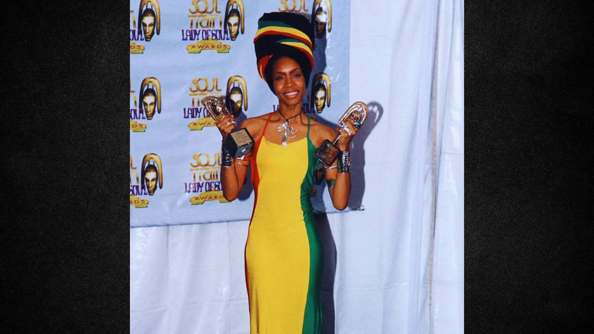 A woman in a yellow and green dress proudly holds two awards in her hands, smiling at the camera.