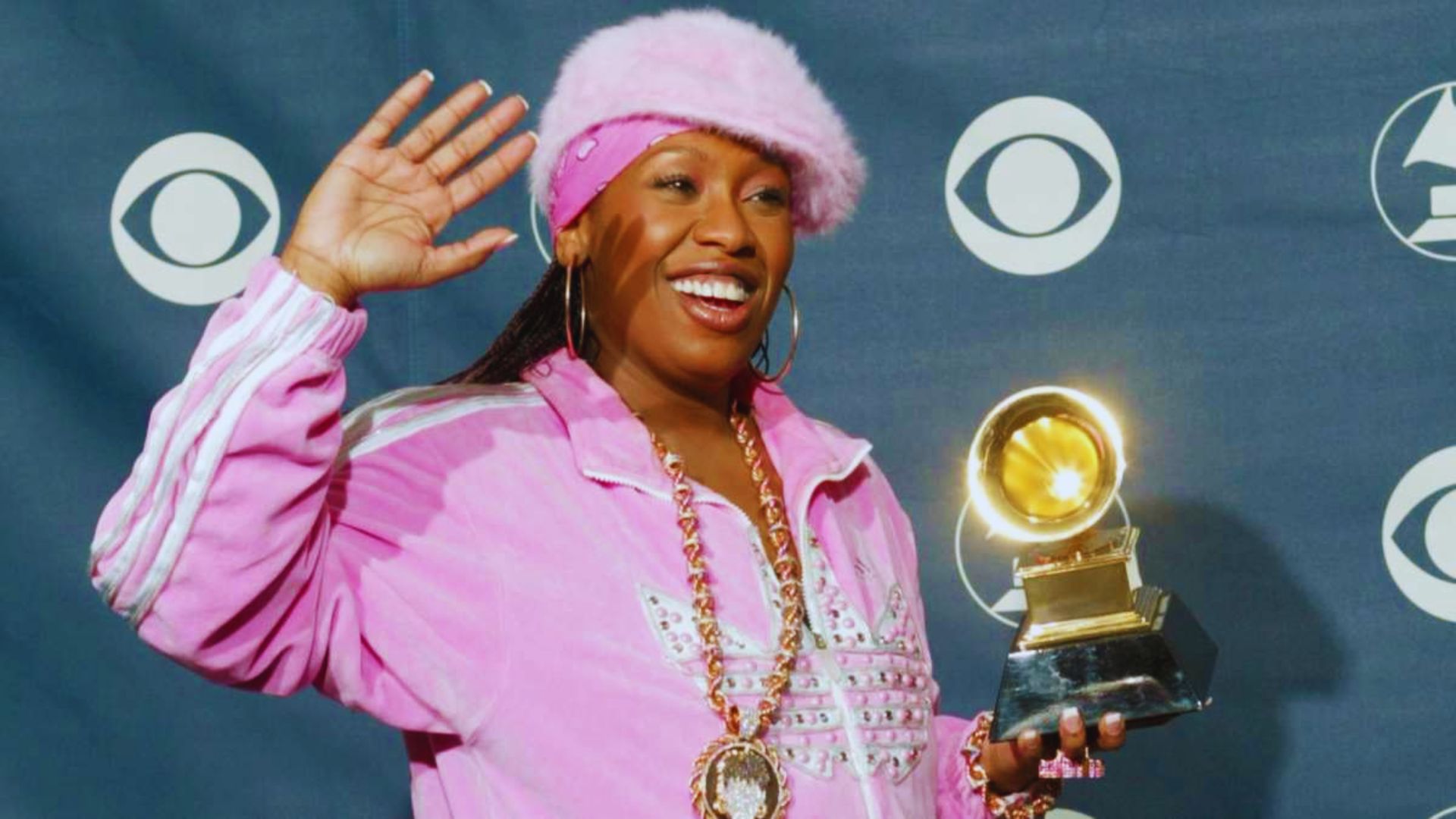 A woman in a pink jacket proudly holds a Grammy Award, smiling at the camera.