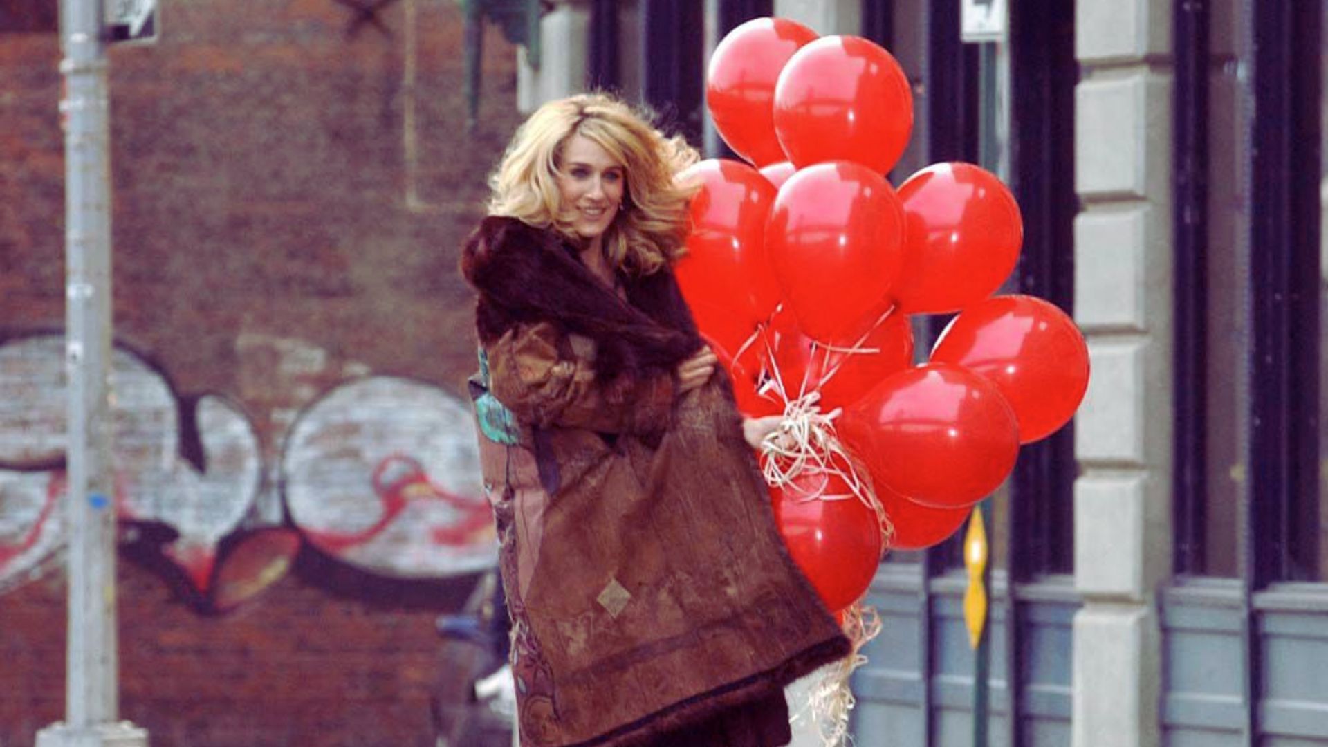 A woman in a coat stands holding a cluster of bright red balloons against a neutral background.
