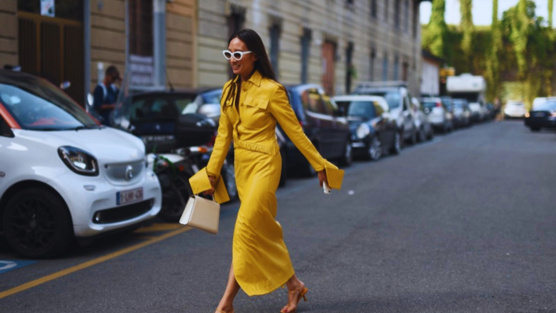 A woman in a bright yellow dress strolls down a city street, surrounded by buildings and greenery.