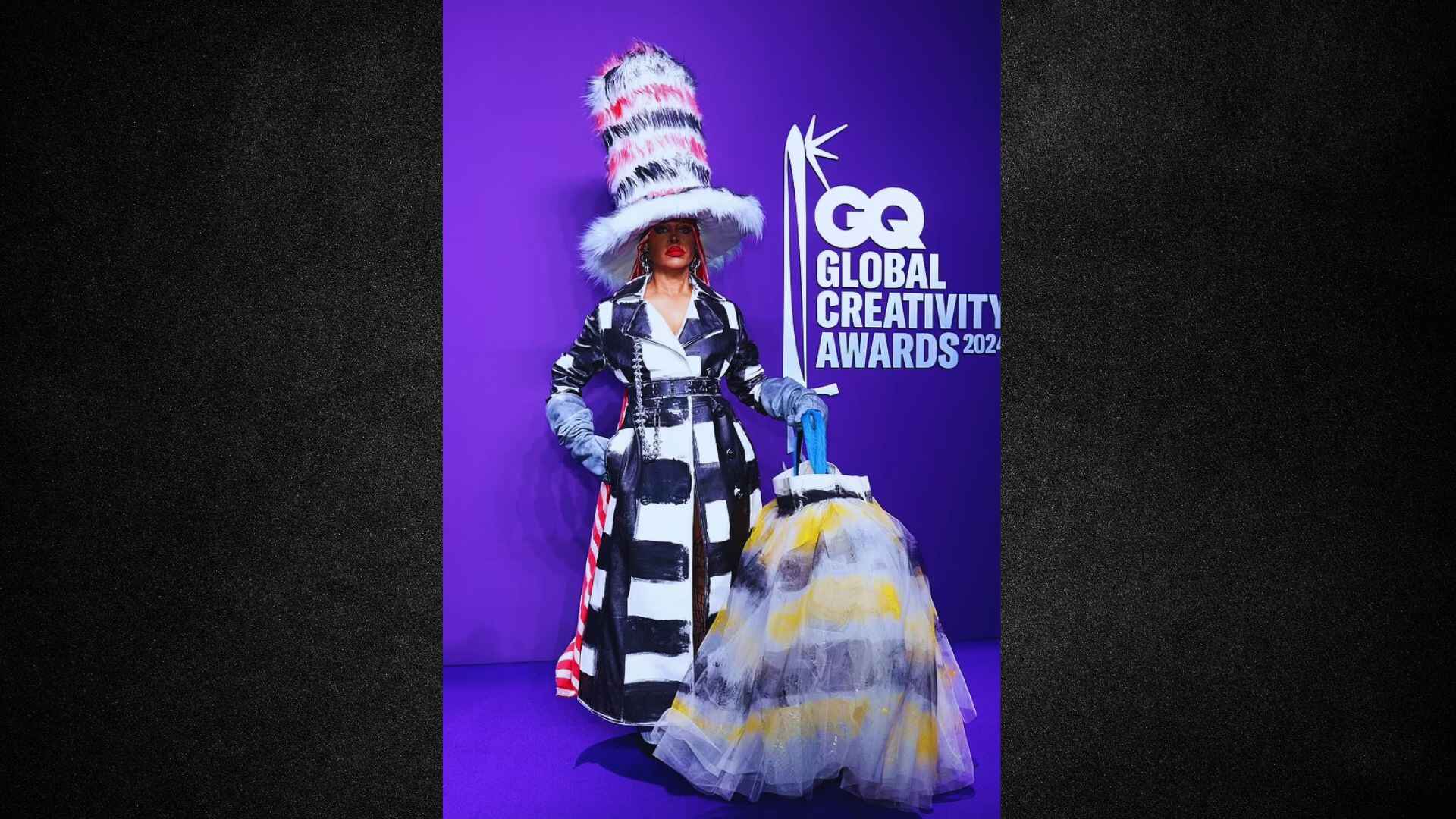 A woman dressed in a costume, wearing a hat and holding a broom, stands in a playful pose.