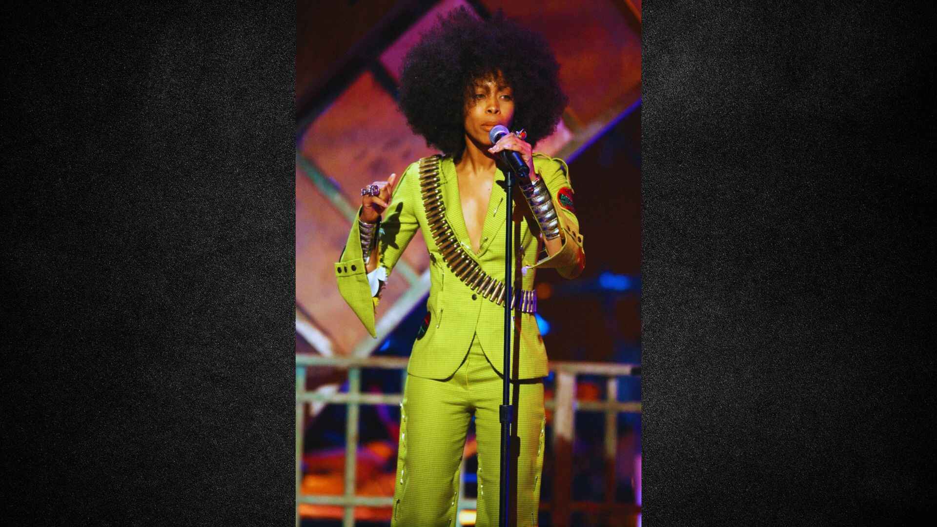 A woman in a green suit stands confidently with a microphone, ready to speak or perform.