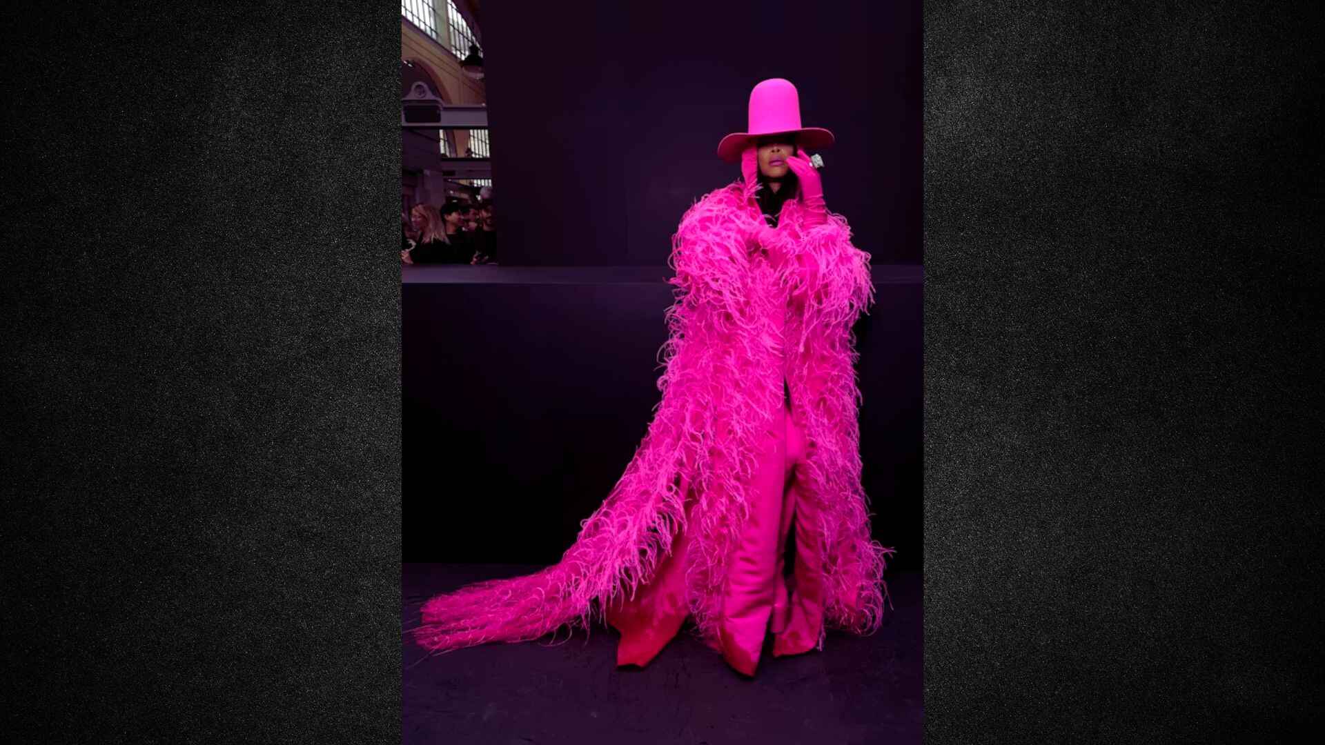 A stylish woman dressed in a pink feather coat and hat, showcasing a bold fashion statement.
