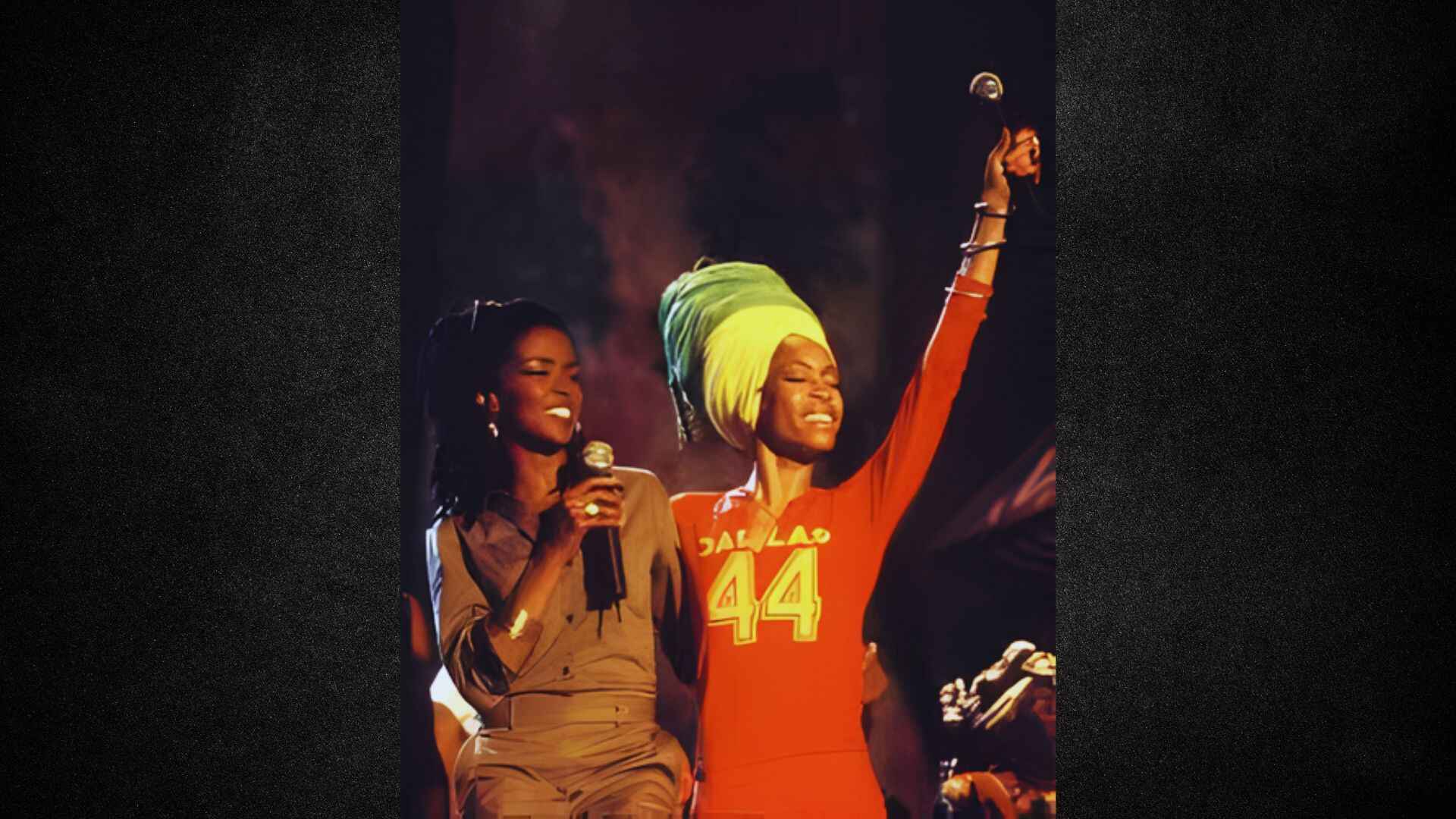 Two women in green shirts and hats are joyfully singing together.