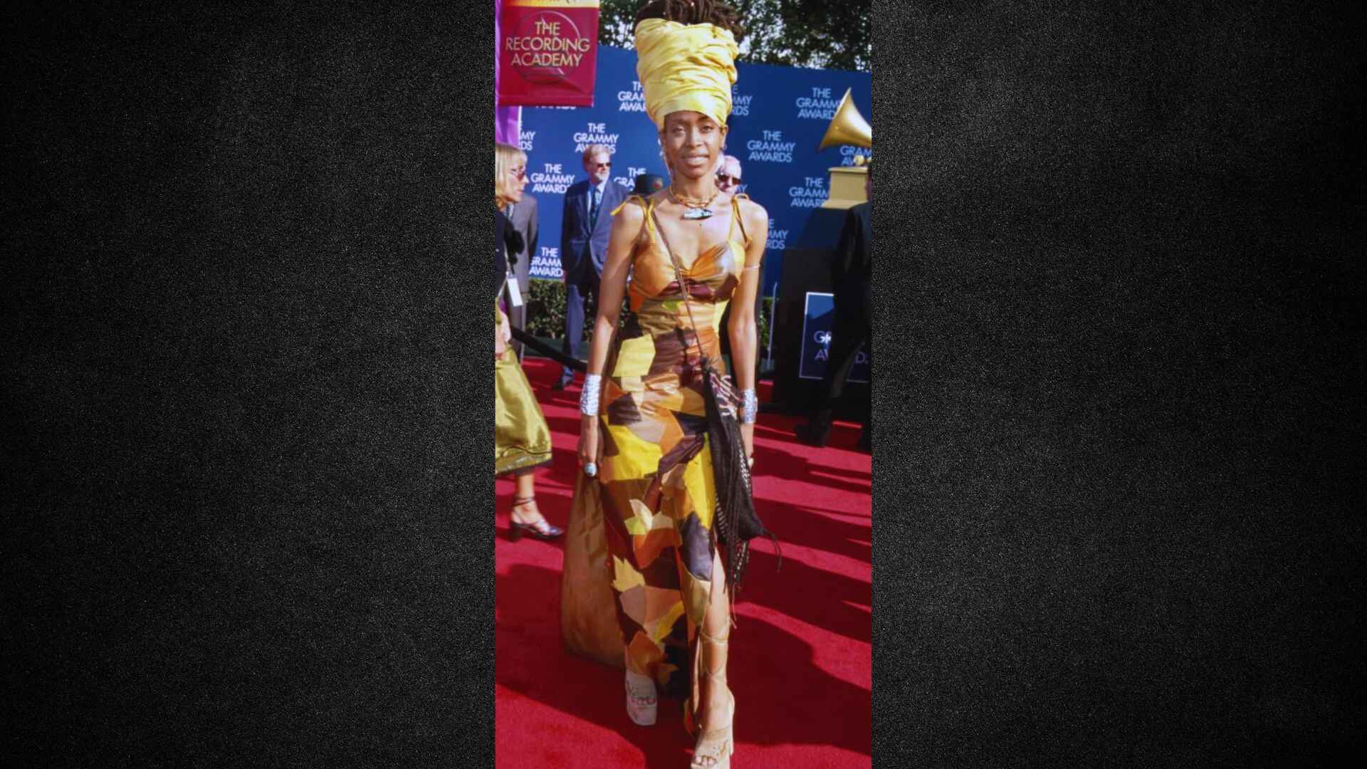A woman in a yellow dress and hat walks confidently on a vibrant red carpet.
