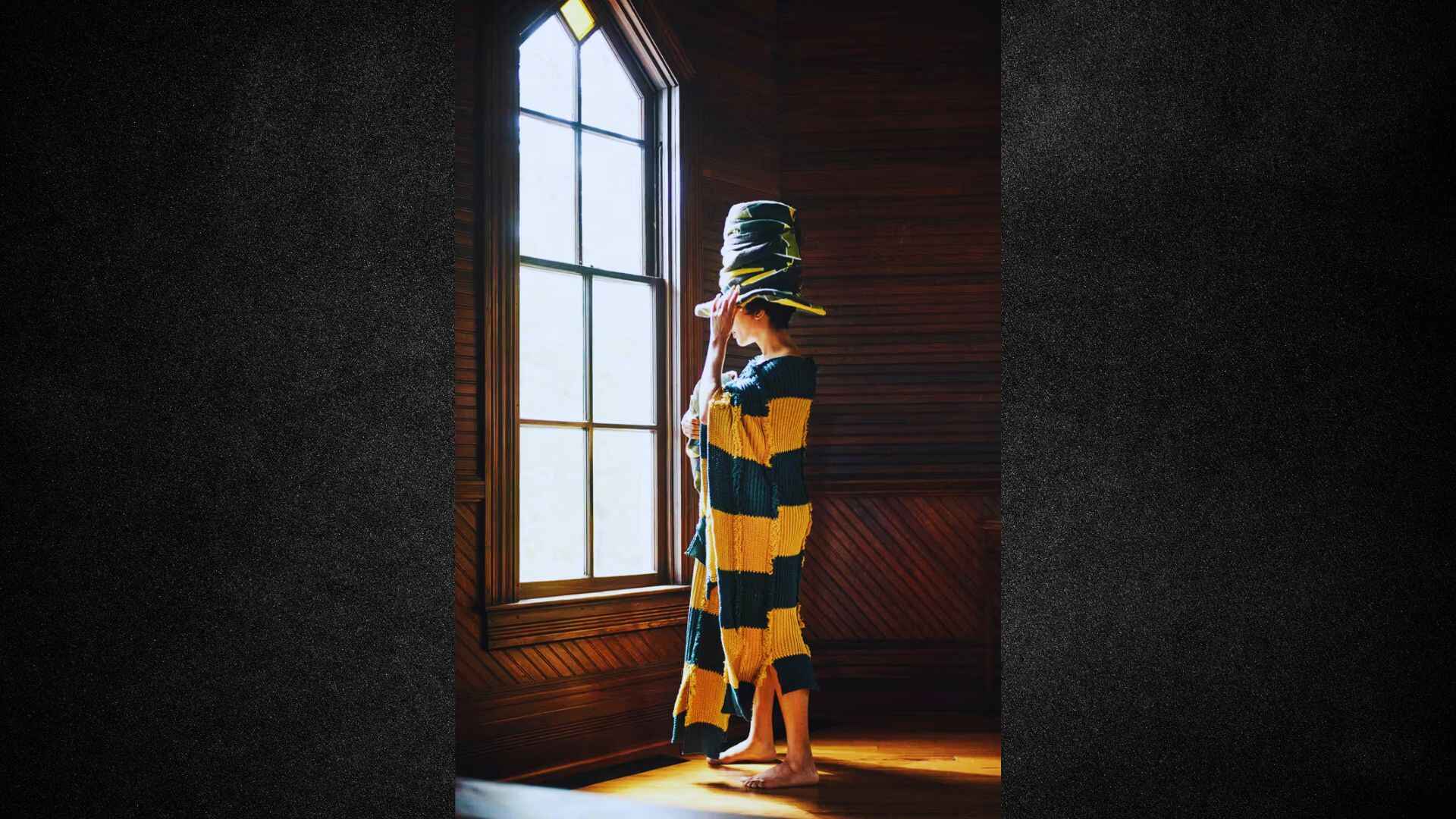 A person stands by a window, wearing a yellow and black striped dress, with natural light illuminating the scene.