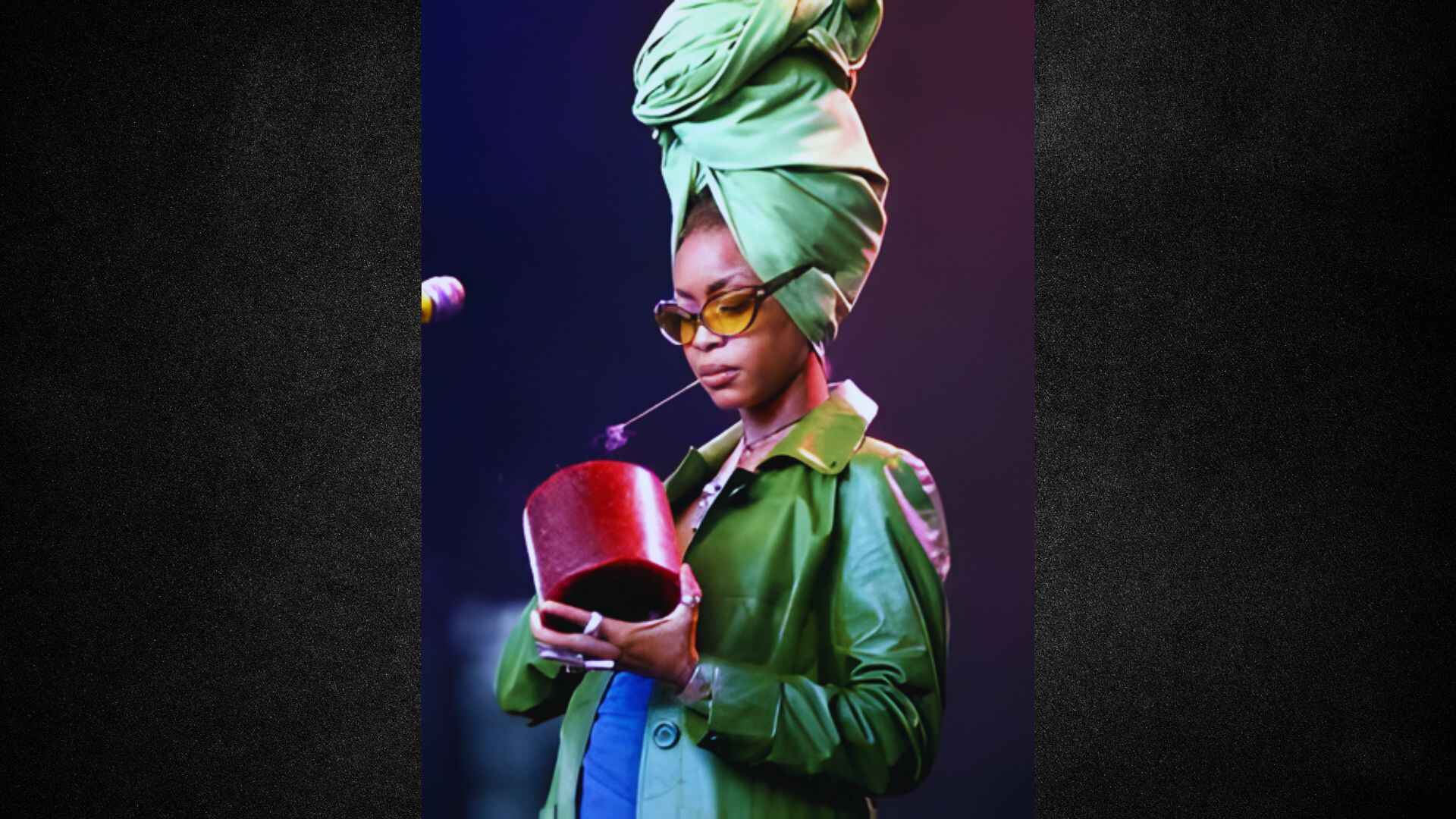 A woman wearing a green jacket and sunglasses holds a red cup, smiling outdoors.
