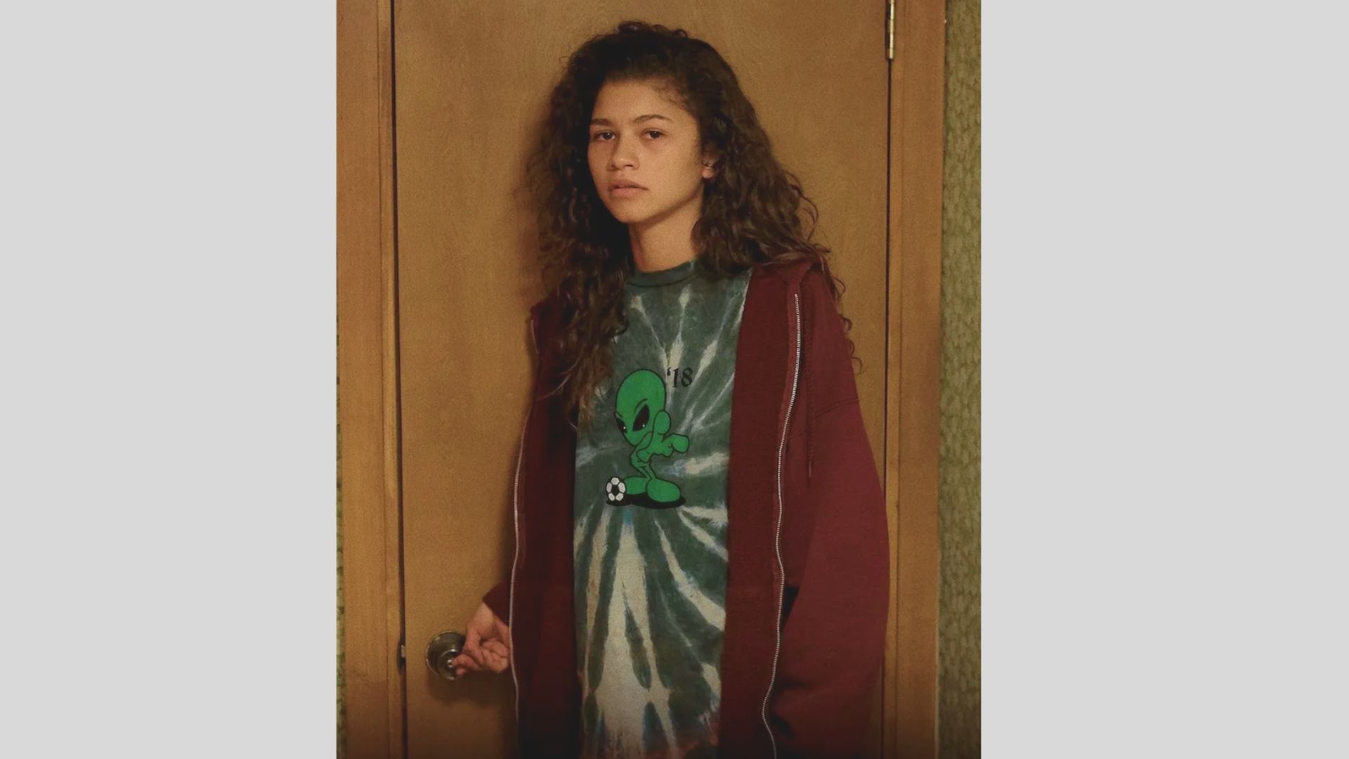 A woman wearing a tie-dye shirt and hoodie stands in front of a closed door, smiling and looking towards the camera.