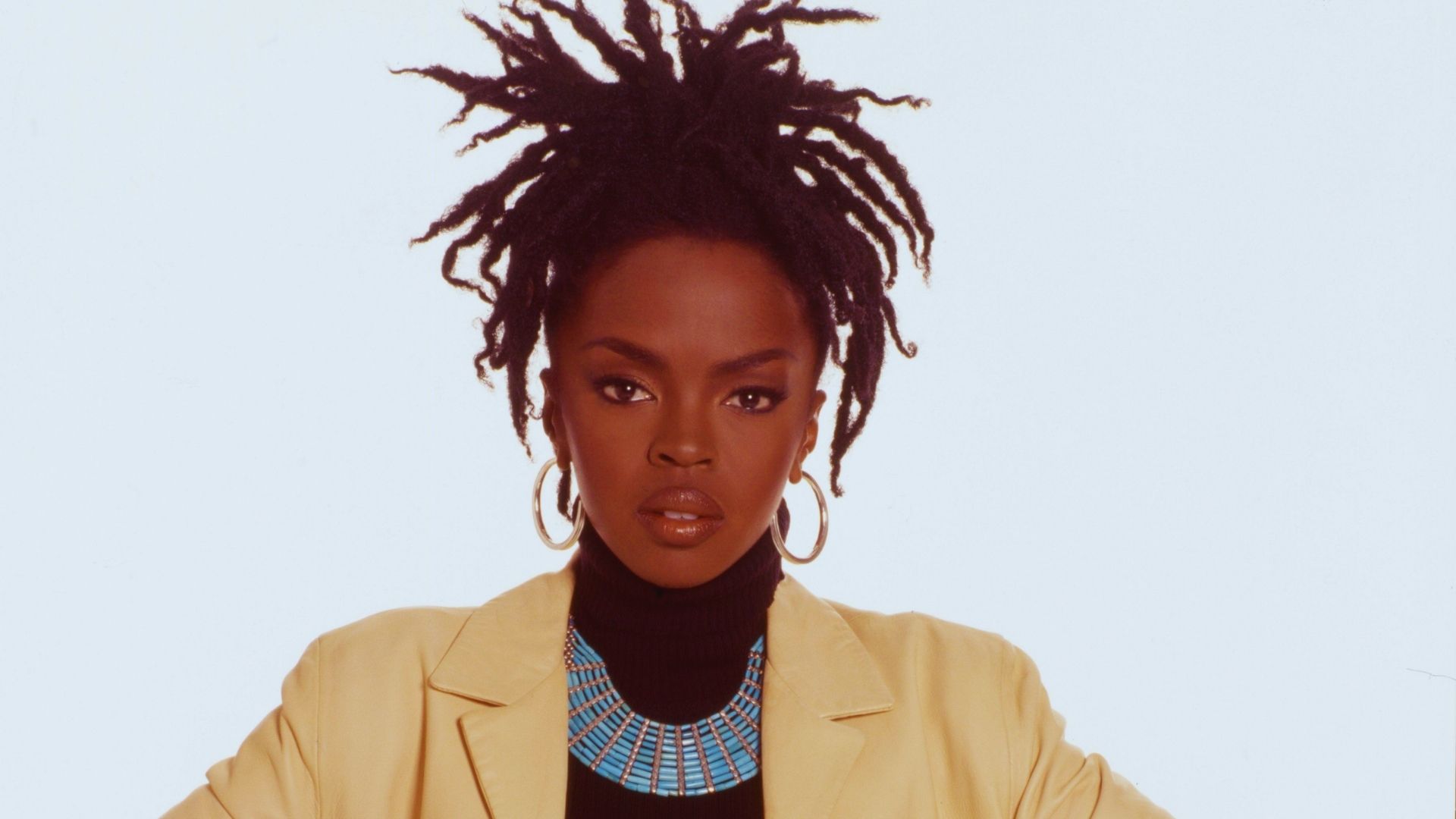 A woman with dreadlocks wearing a bright yellow jacket stands confidently, showcasing her unique style.