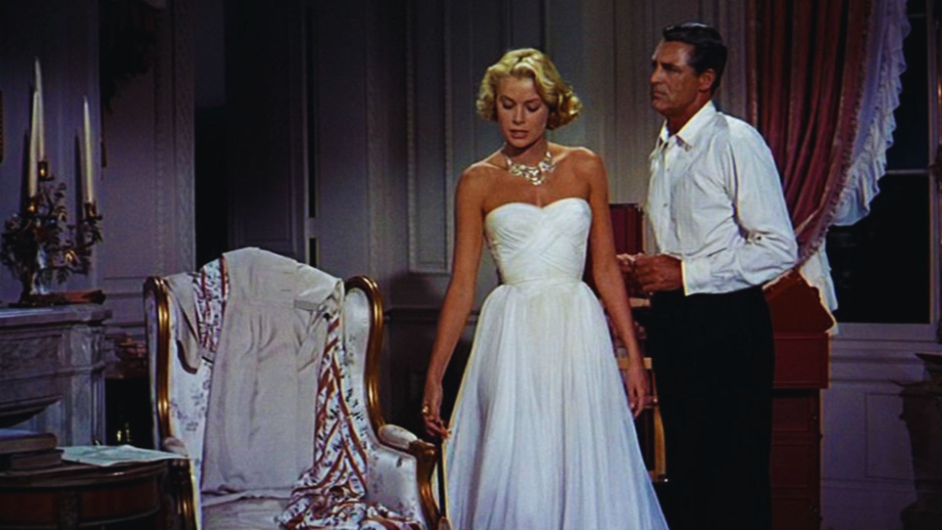A man and woman in elegant white dresses stand side by side, smiling at the camera in a bright outdoor setting.