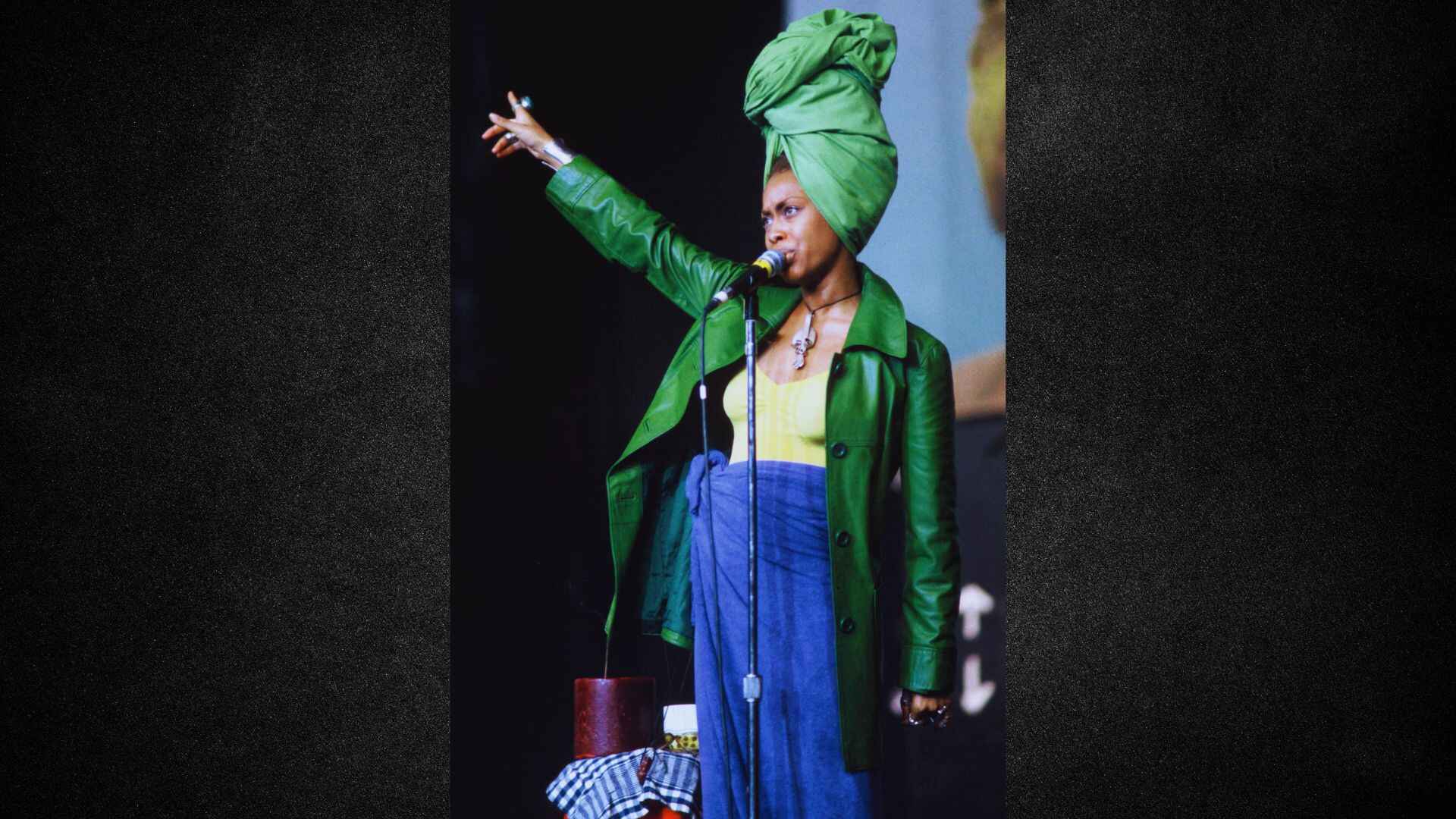 A woman wearing a green turban holds a microphone, ready to speak or perform.