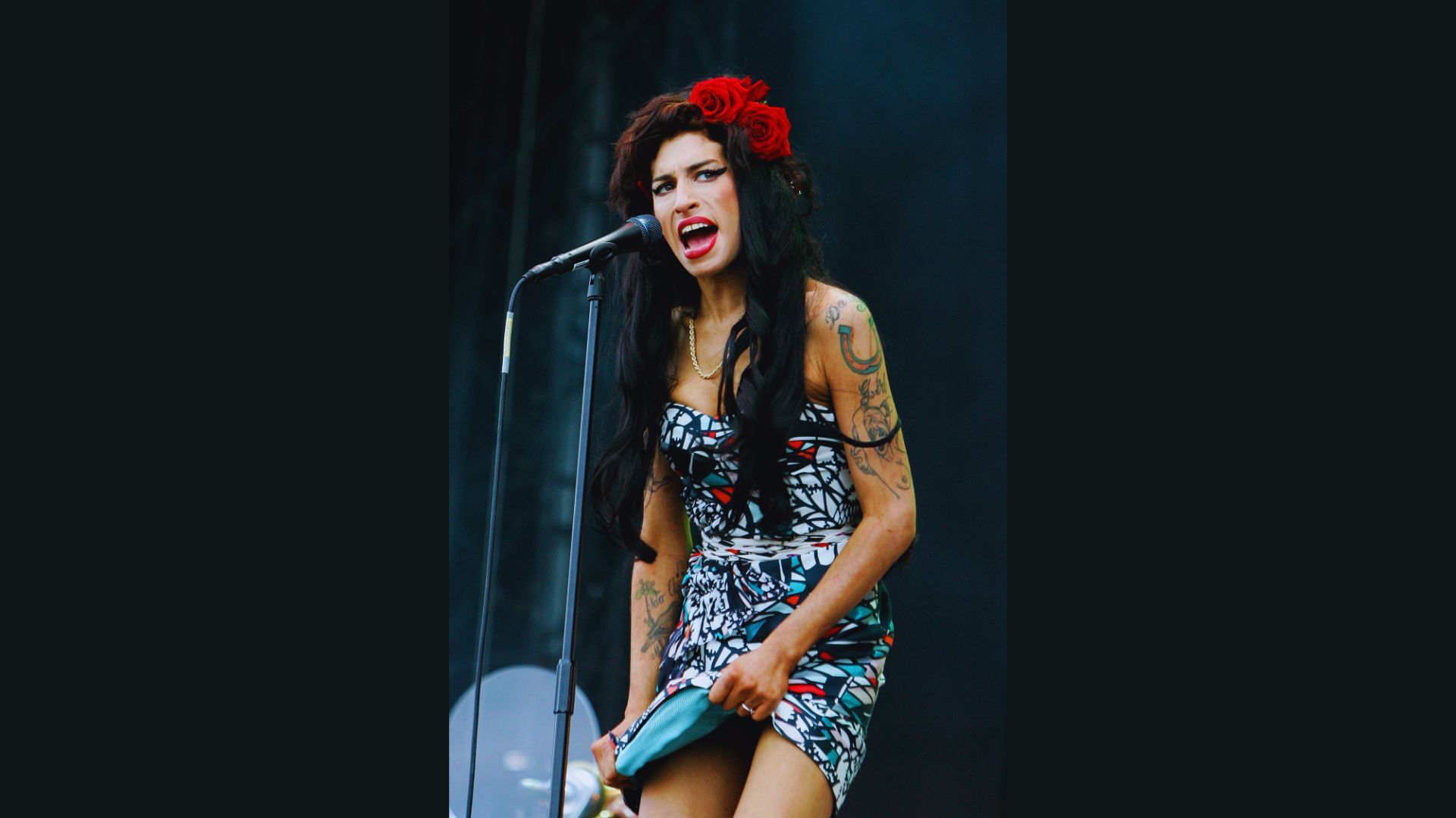 A woman with leg tattoos holds a microphone, poised to speak or perform, showcasing her unique style and confidence.