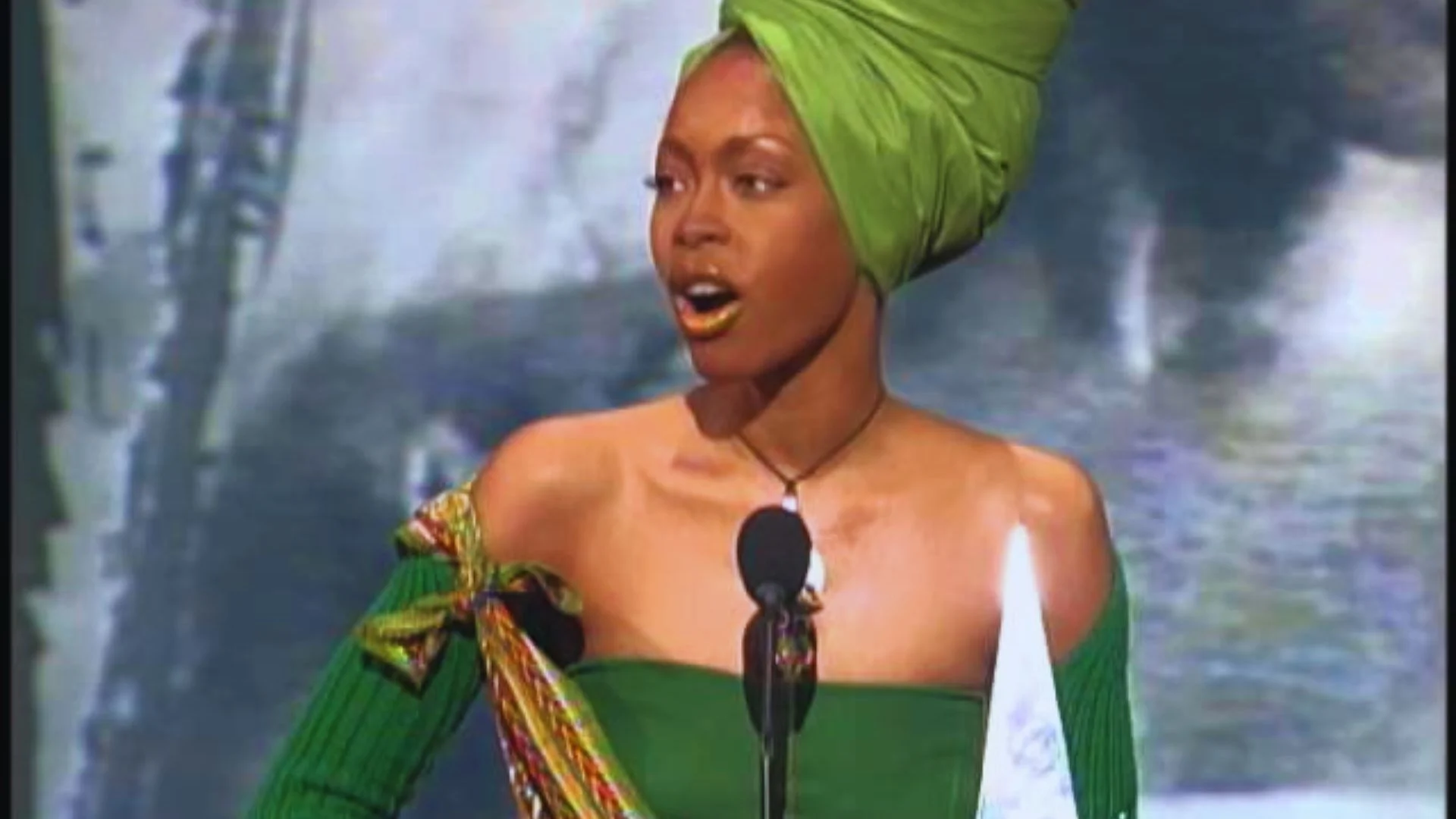 A woman in a green dress and turban speaks into a microphone during a public event.