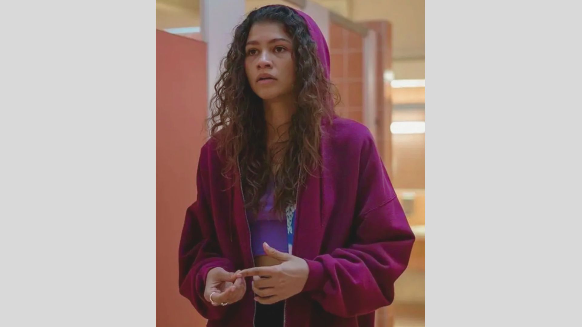 A woman wearing a purple sweater stands in a hallway, with walls and floor visible in the background.