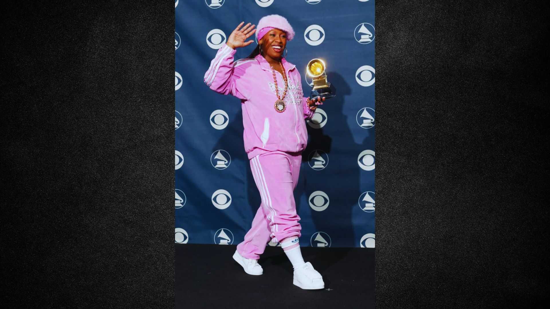 A man in a pink suit and white sneakers proudly holds a trophy, smiling at the camera.