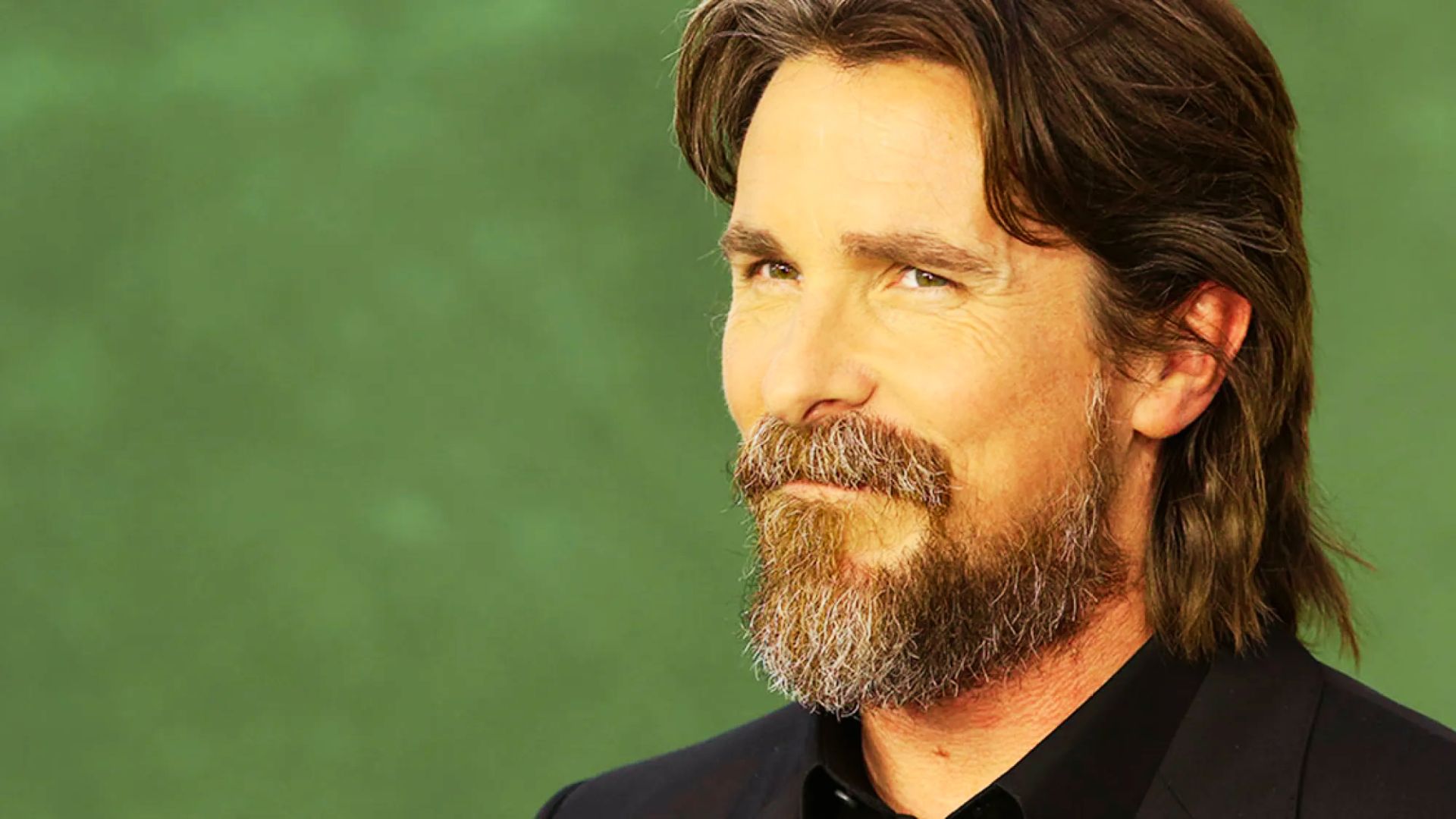 Christian Bale with long hair and a beard, dressed in a suit.