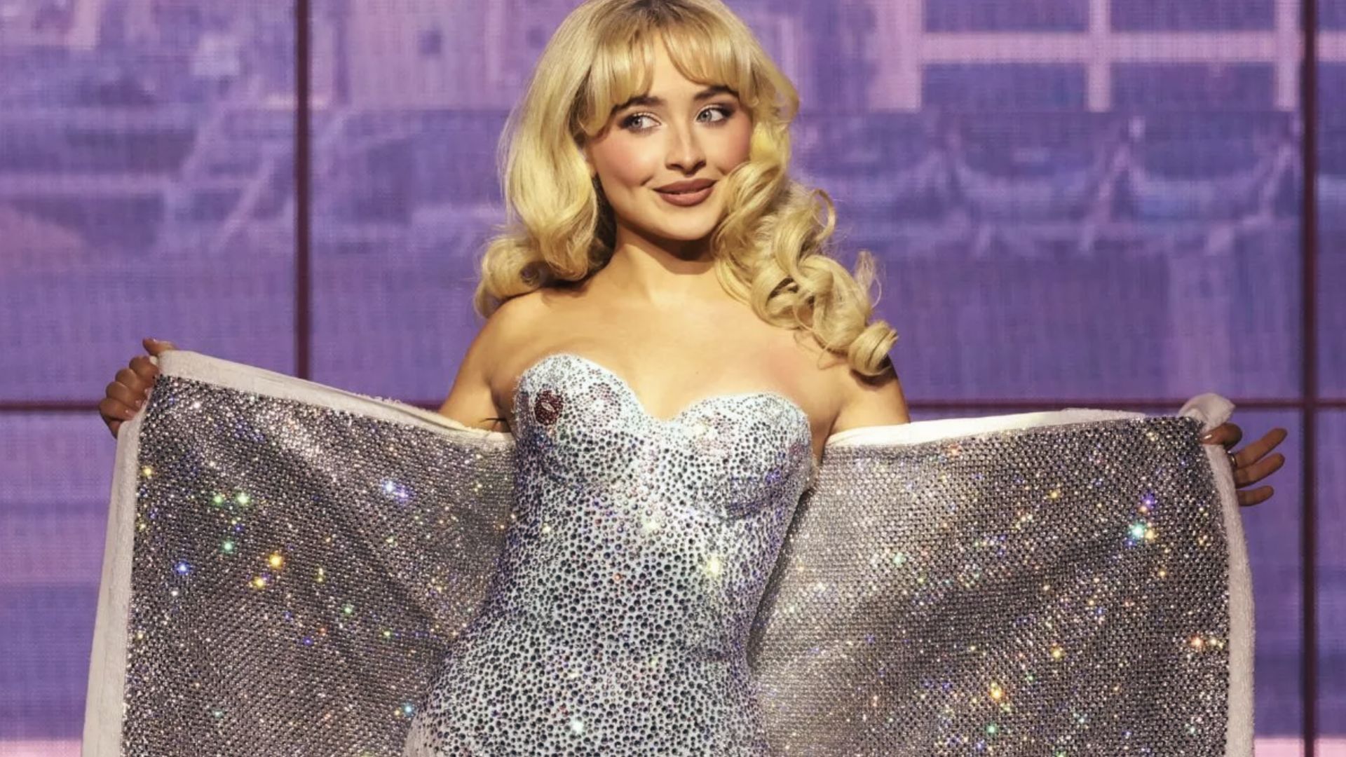 Sabrina Carpenter holds open a rhinestone towel while wearing a strapless silver sparkly mini dress.