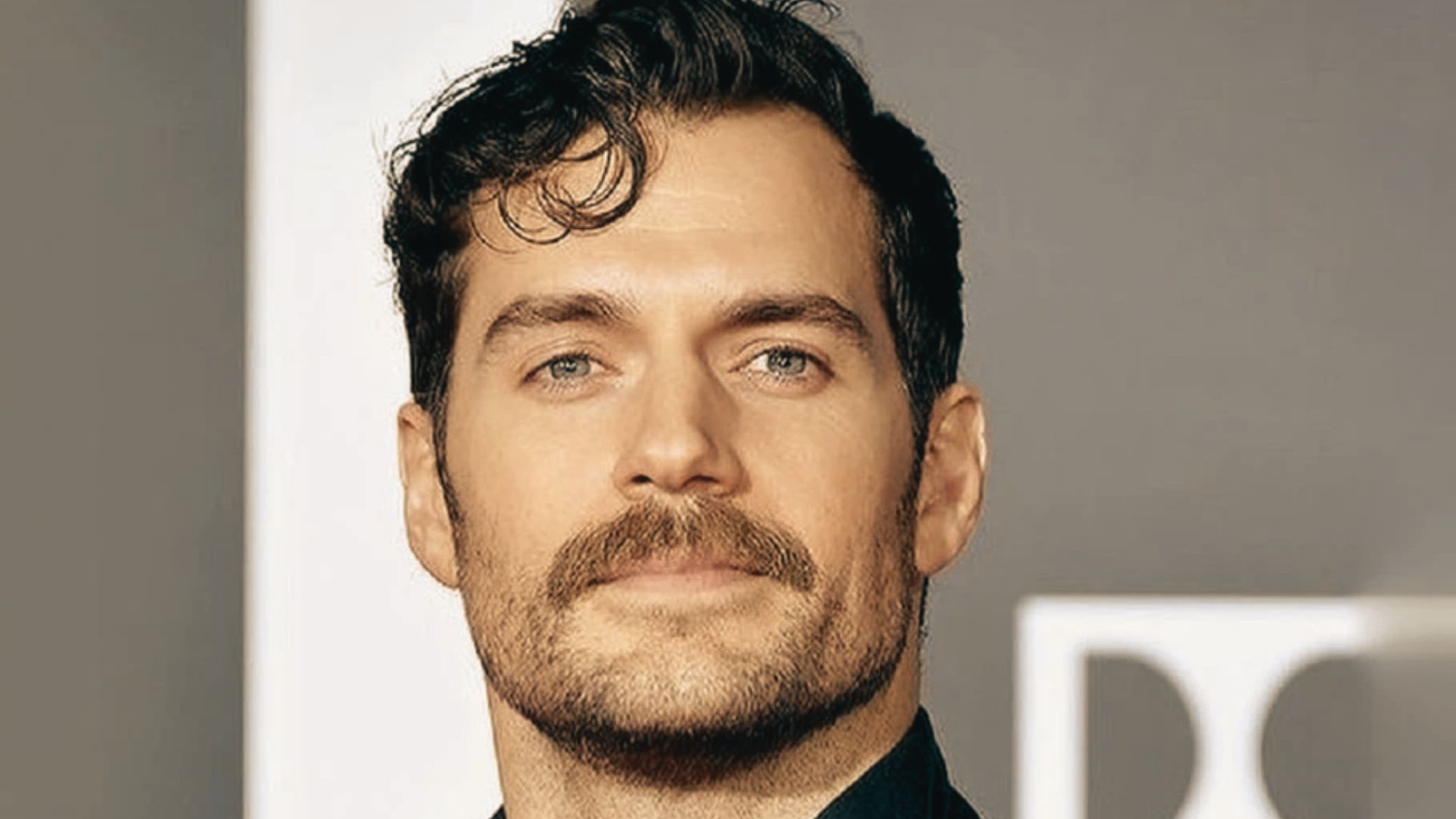 Henry Cavill with a mustache wearing a formal suit, standing confidently with a neutral expression.