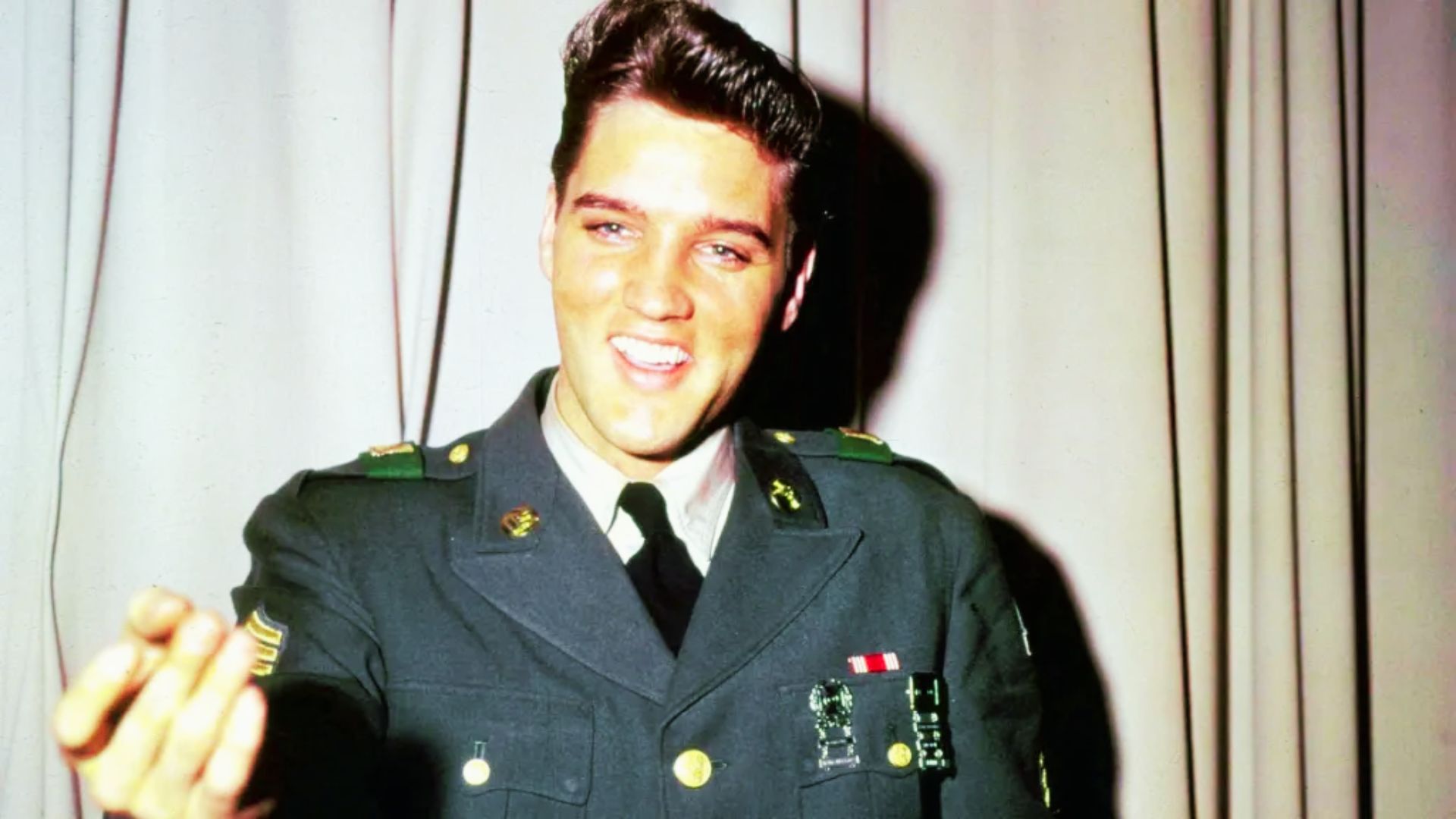Elvis Presley in military uniform, hand outstretched, showcasing his iconic hairstyle during his Army service.